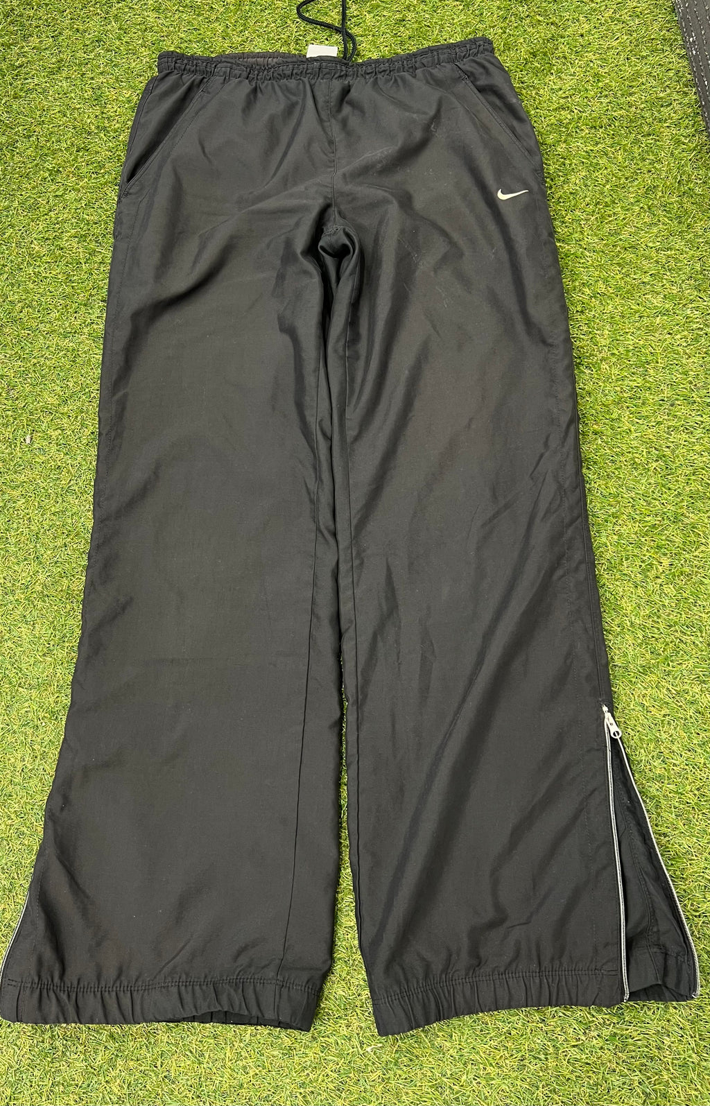 Front view of Nike black loose fit track pants with elastic waistband and zipped side ankle detail on grass surface