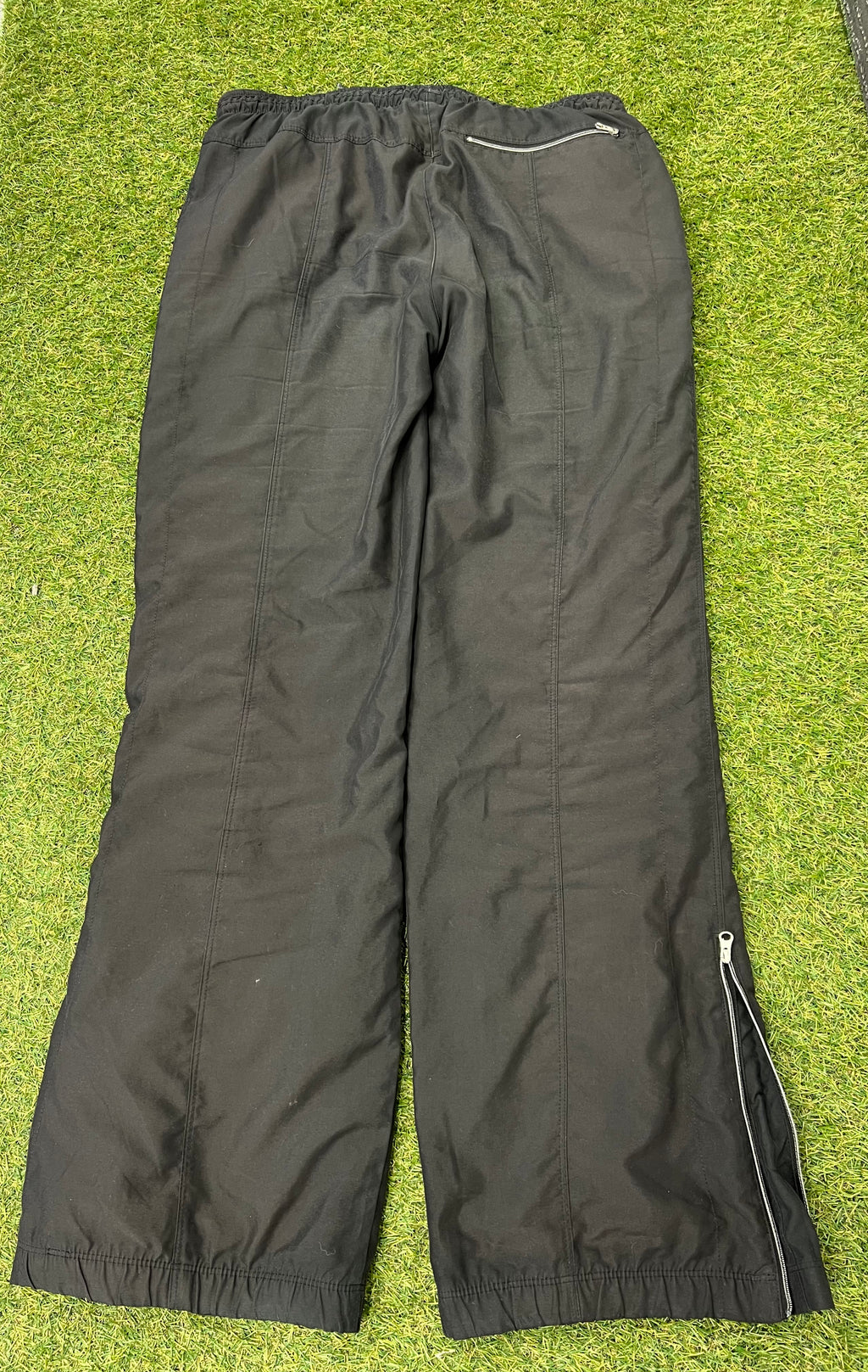 Rear view of Nike black loose fit track pants showing back zip pocket and side zipped ankle openings on grass