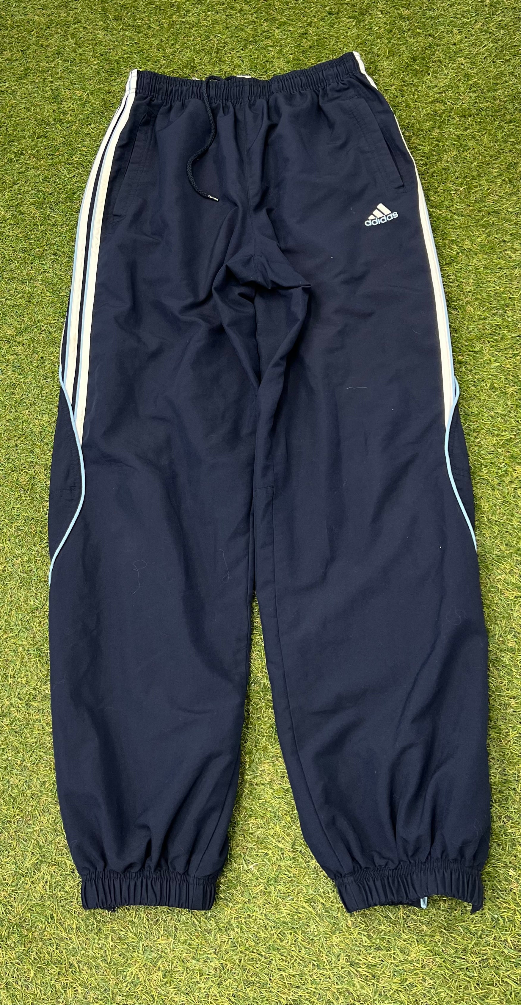 Front view of men's Adidas navy tracksuit bottoms with white side stripes and elasticated ankles laid flat on grass