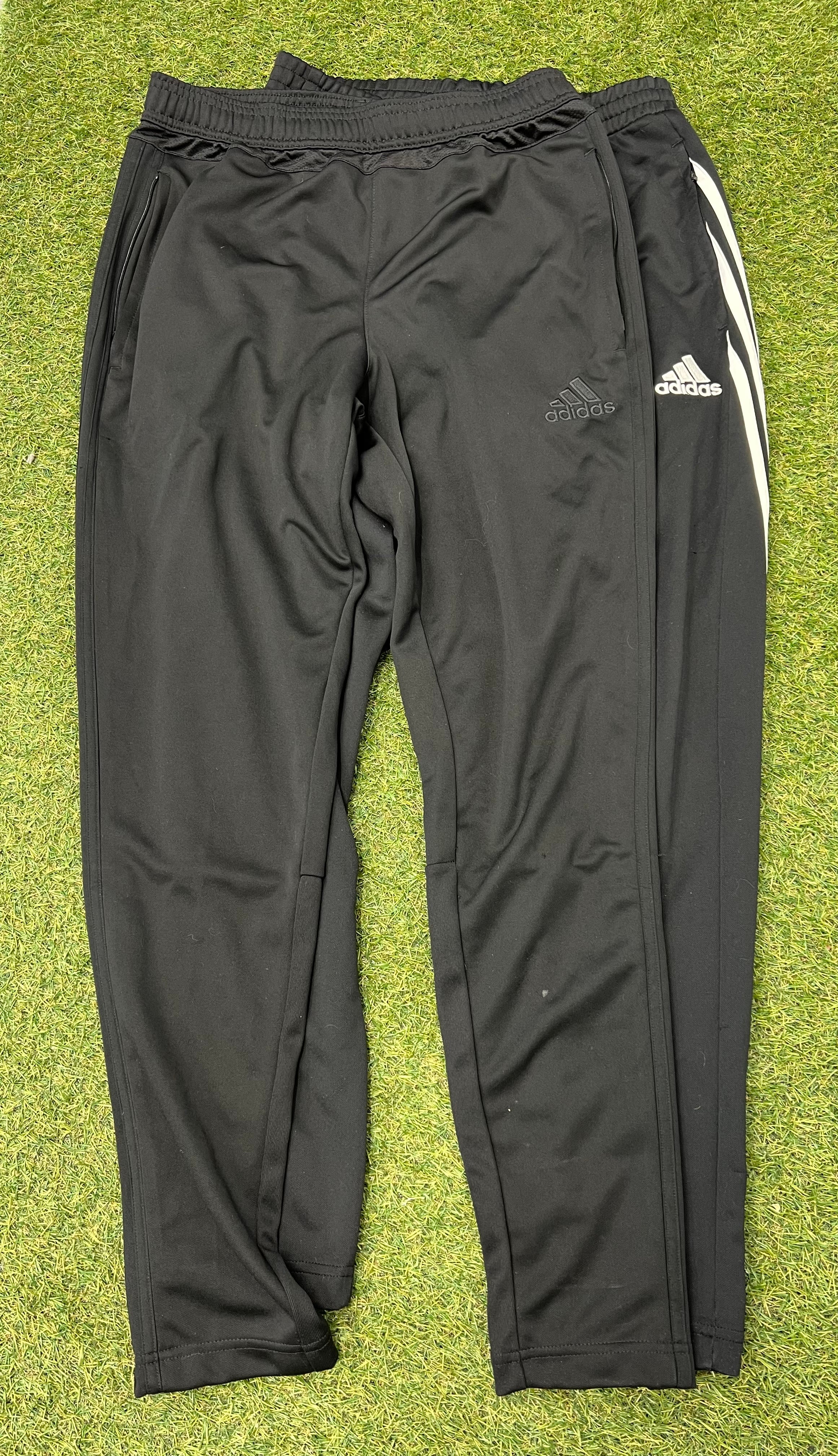 Pair of Adidas black tracksuit bottoms laid flat on grass, front view with elastic waistband visible