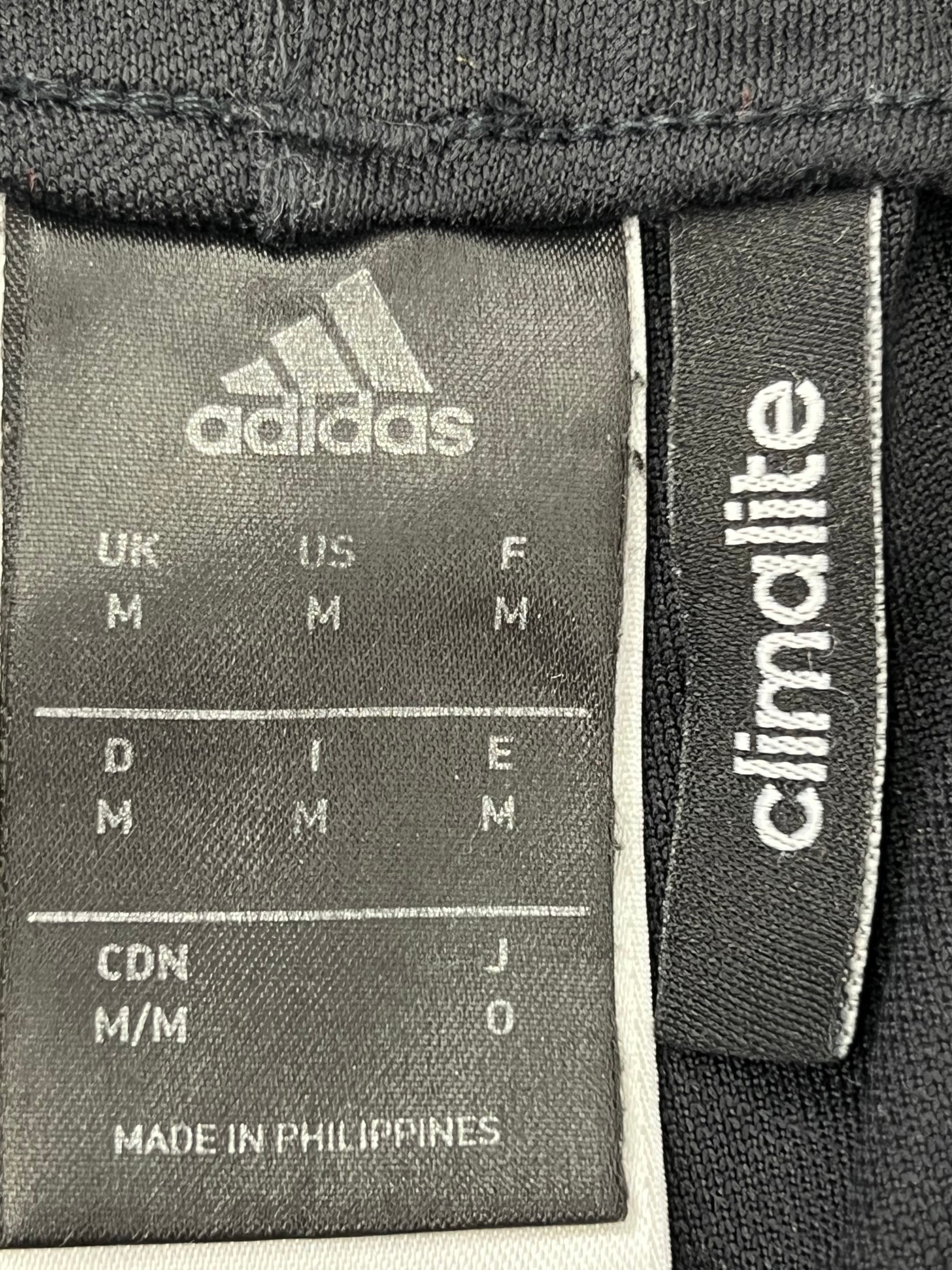 Front view of Adidas black tracksuit bottoms with tapered legs and ankle zippers open on grass