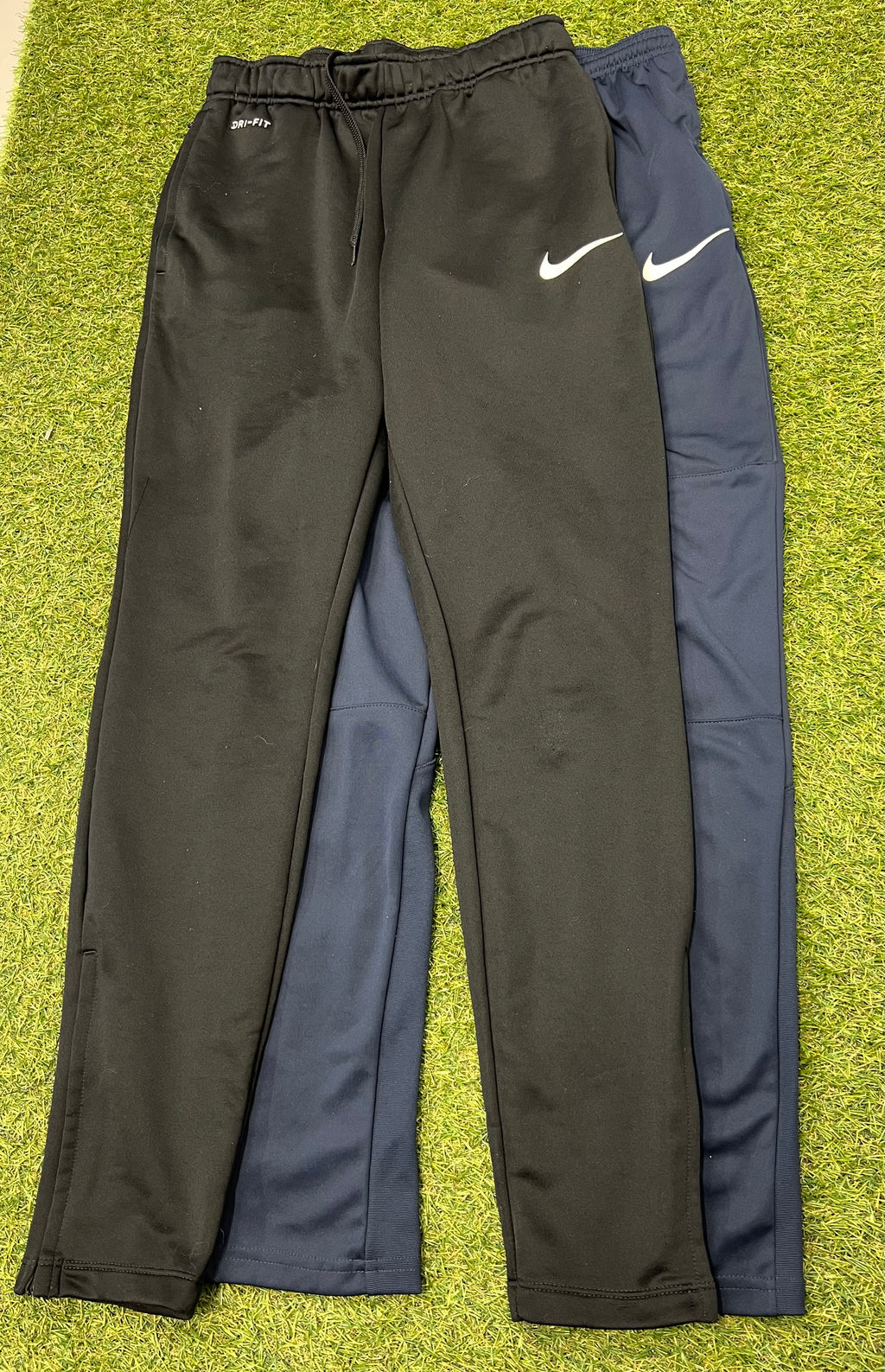 Paired Nike Dri-FIT tracksuit bottoms in black and navy laid flat on grass