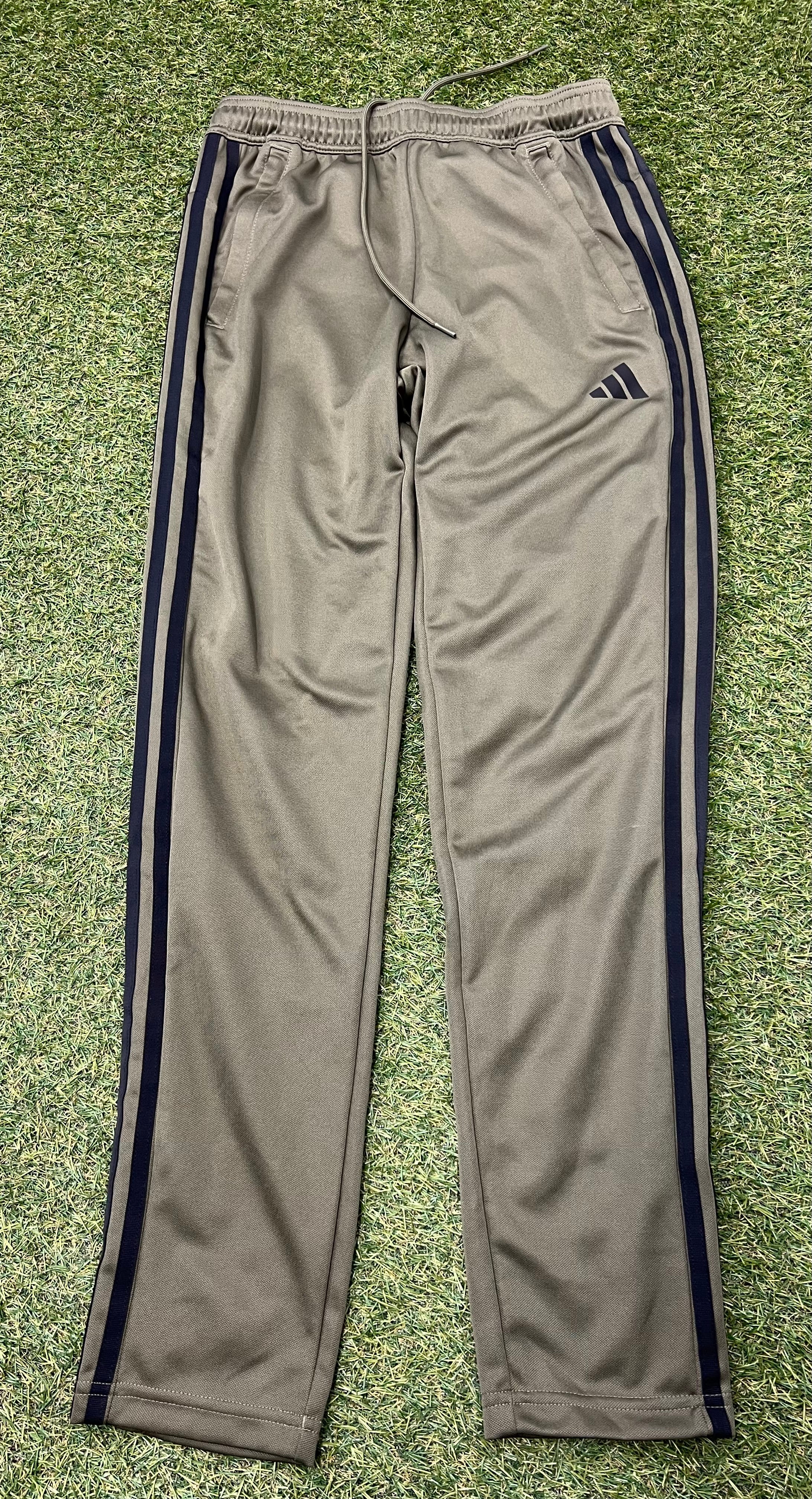 Front view of men's khaki tracksuit bottoms with black side stripes and an elastic waistband with drawstring, laid flat on grass.