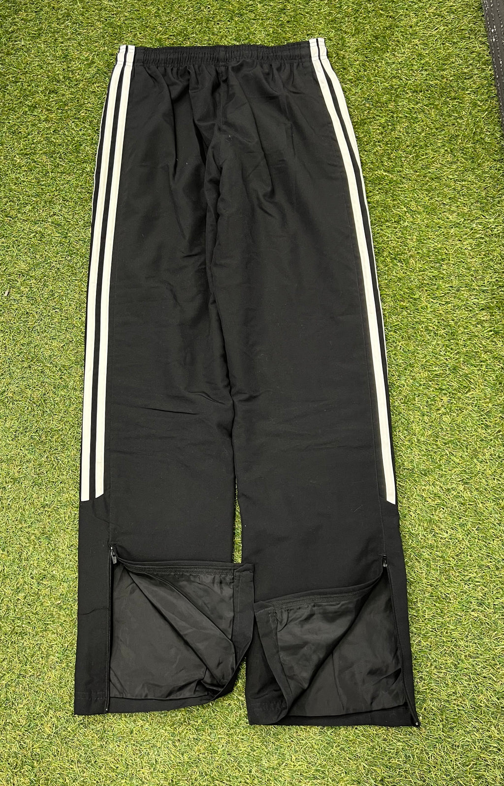 Back view of black adidas women's tracksuit bottoms showing elastic waistband and ankle zips on grass background