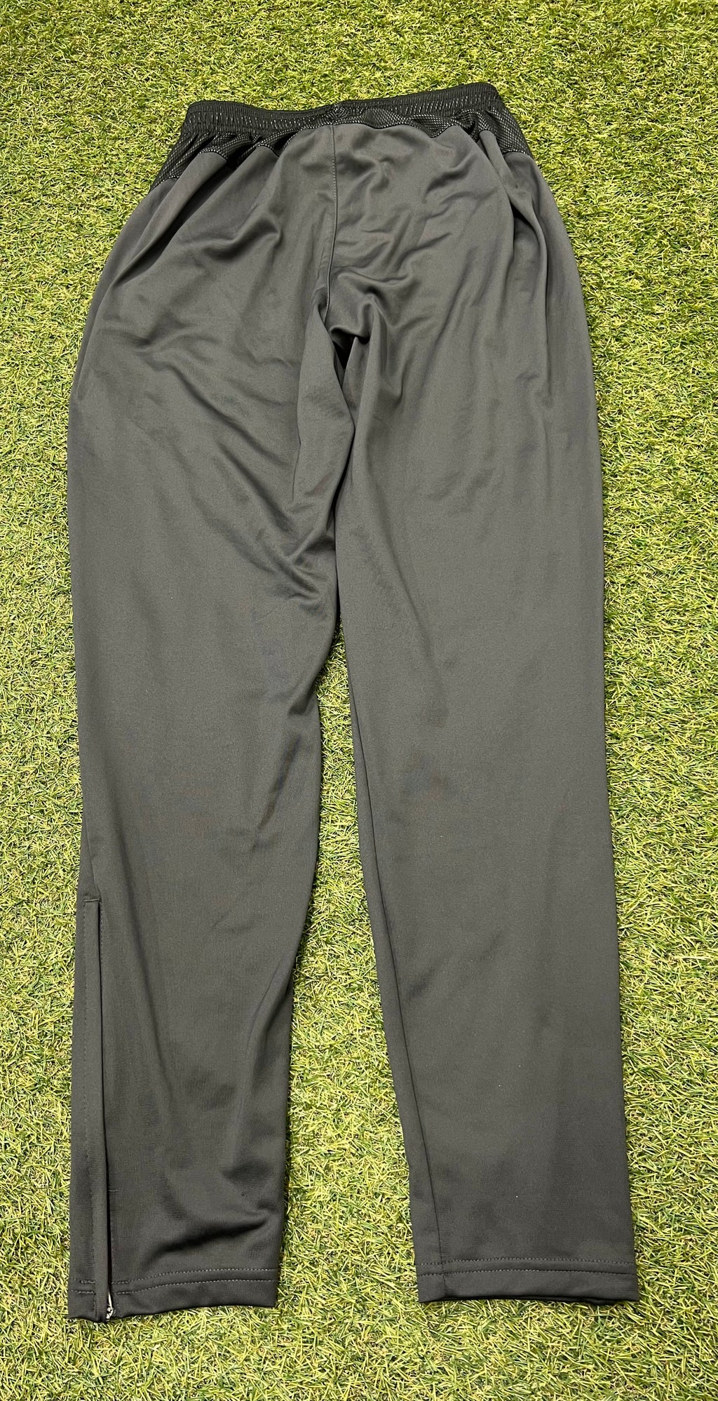 Back view of men's Nike Dri-FIT charcoal tracksuit trousers laid flat on grass showing smooth fabric and elastic waistband