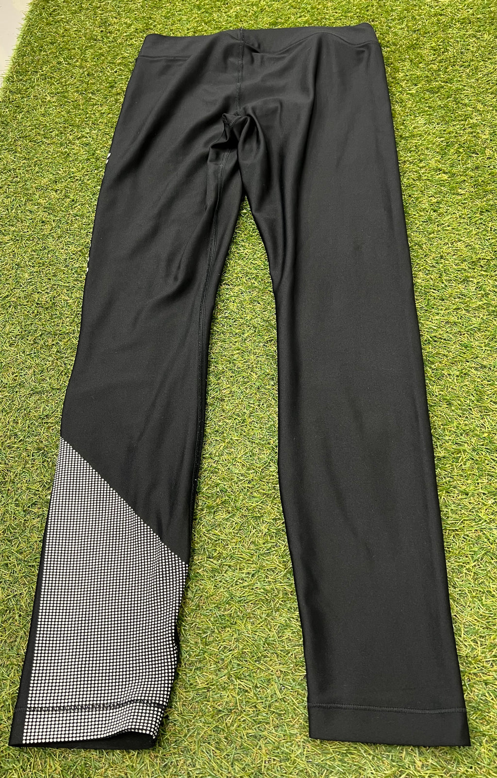 Back view of black Under Armour compression tights showing reflective patterned detail on lower left leg, laid flat on grass