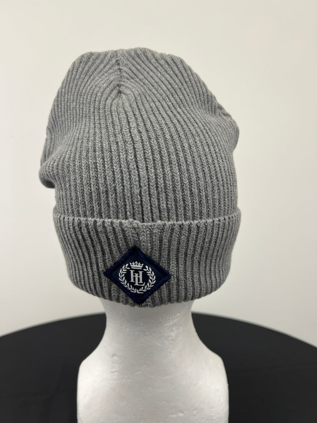 Back view of grey ribbed knit beanie hat with navy Henri Lloyd embroidered badge on cuff displayed on a white mannequin head