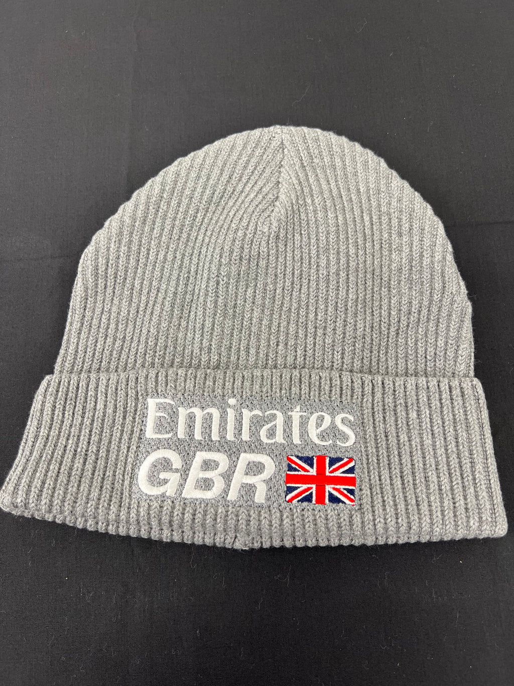 Flat lay of grey ribbed knit beanie hat showing white Emirates GBR logo and British flag embroidery on cuff against black background