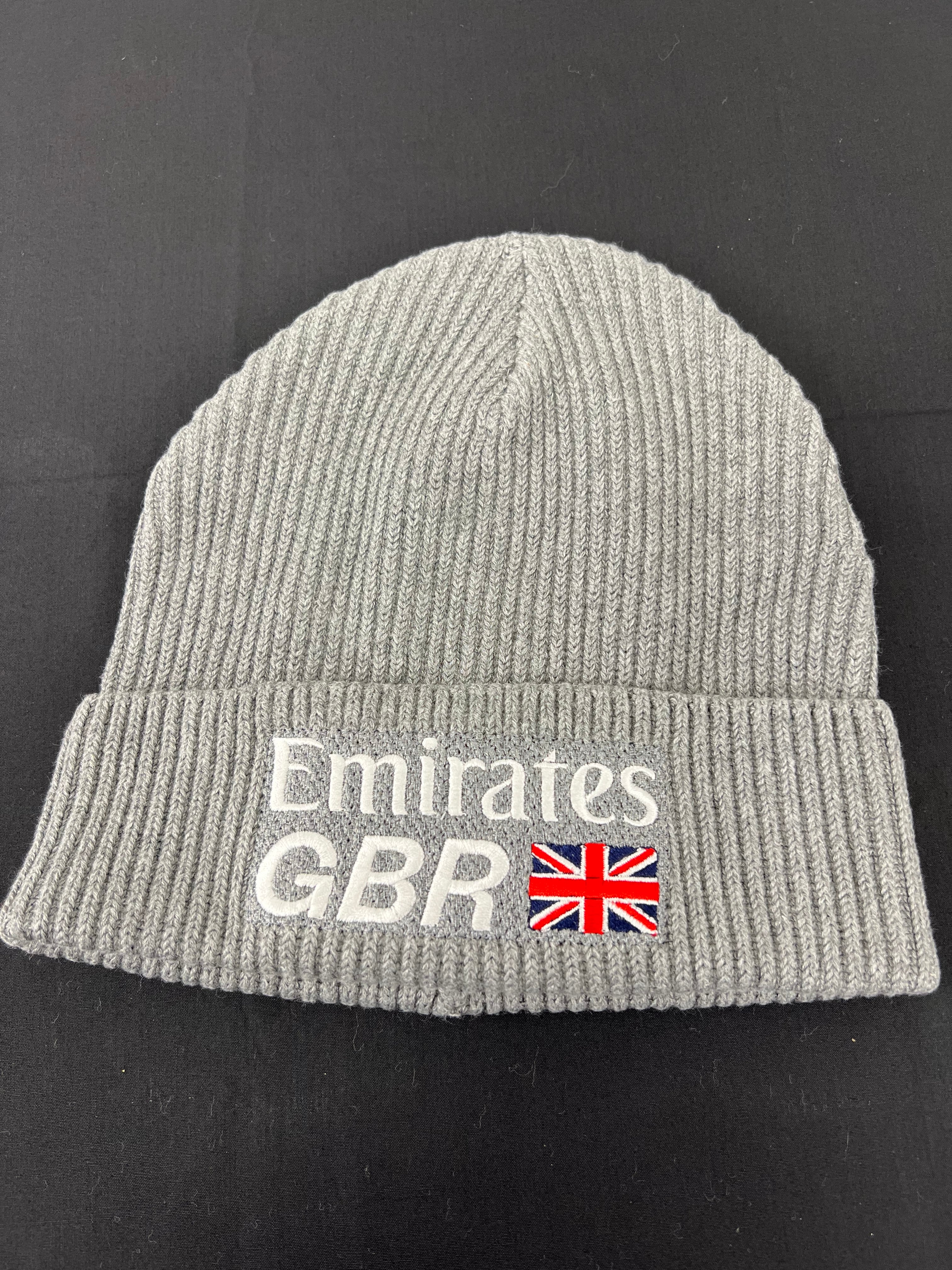 Flat lay of grey ribbed knit beanie hat showing white Emirates GBR logo and British flag embroidery on cuff against black background