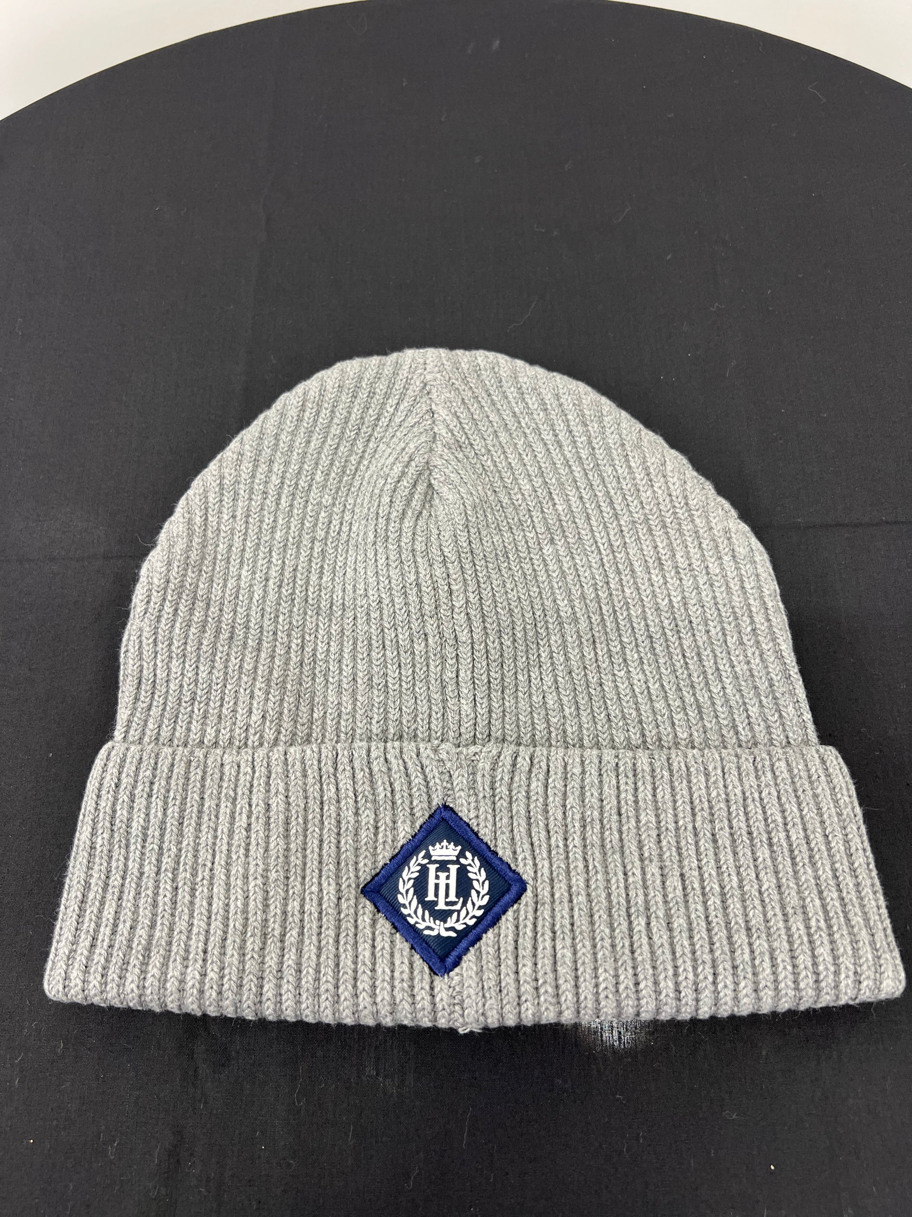 Flat lay of grey ribbed knit beanie hat showing navy Henri Lloyd embroidered badge on cuff against black background