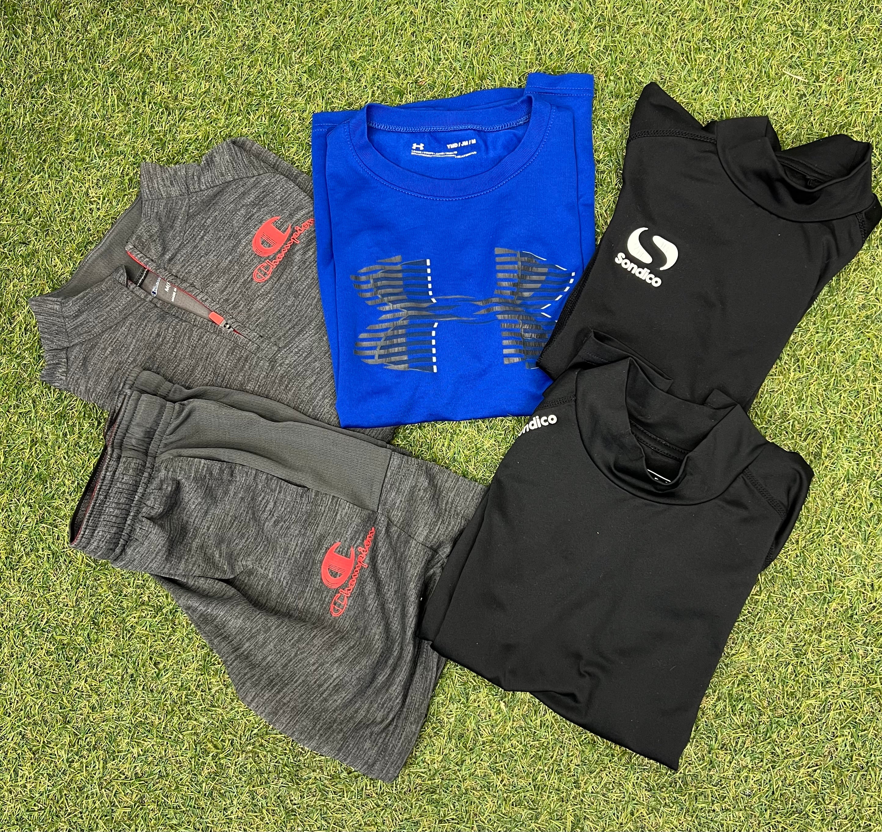 Kids sportswear bundle including grey Champion tracksuit, blue Under Armour T-shirt, and black Sondico tops laid on grass