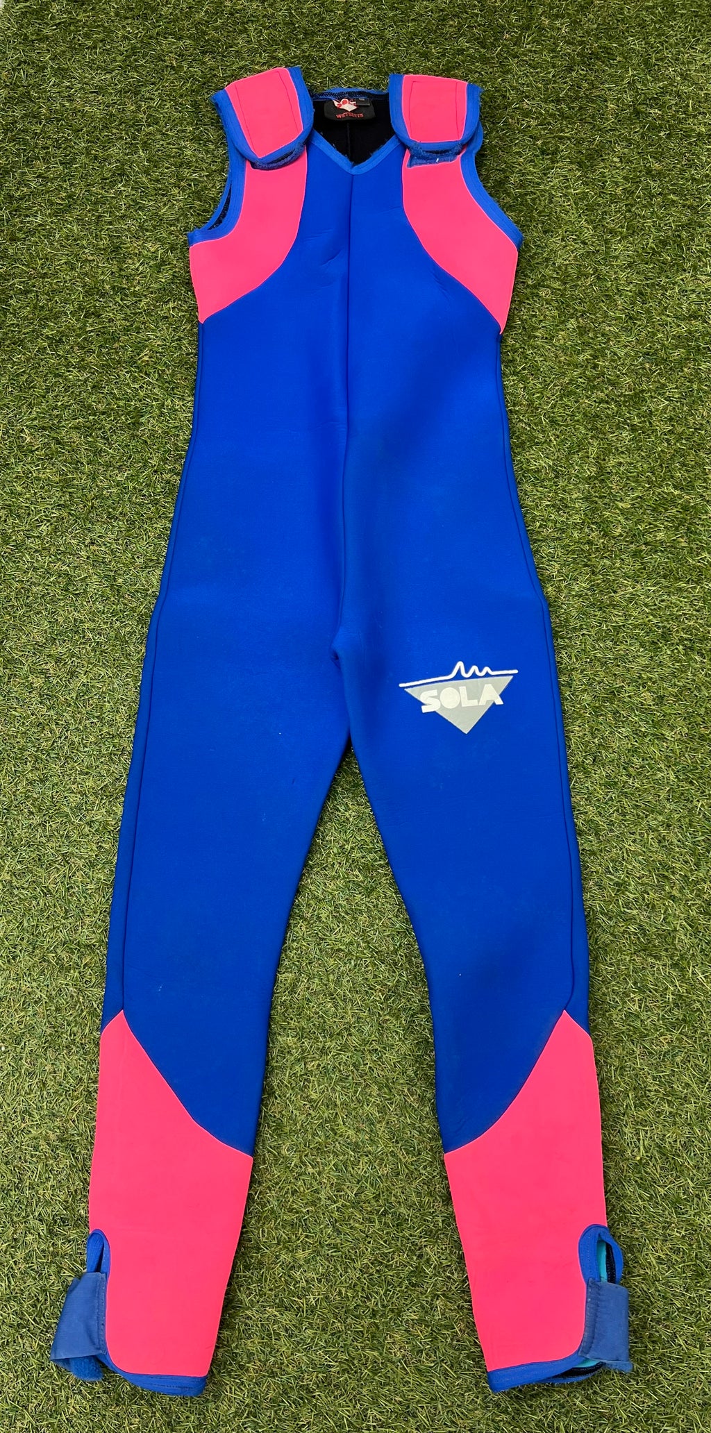 Front view of blue sleeveless wetsuit with bright pink panels on shoulders and lower legs laid on grass