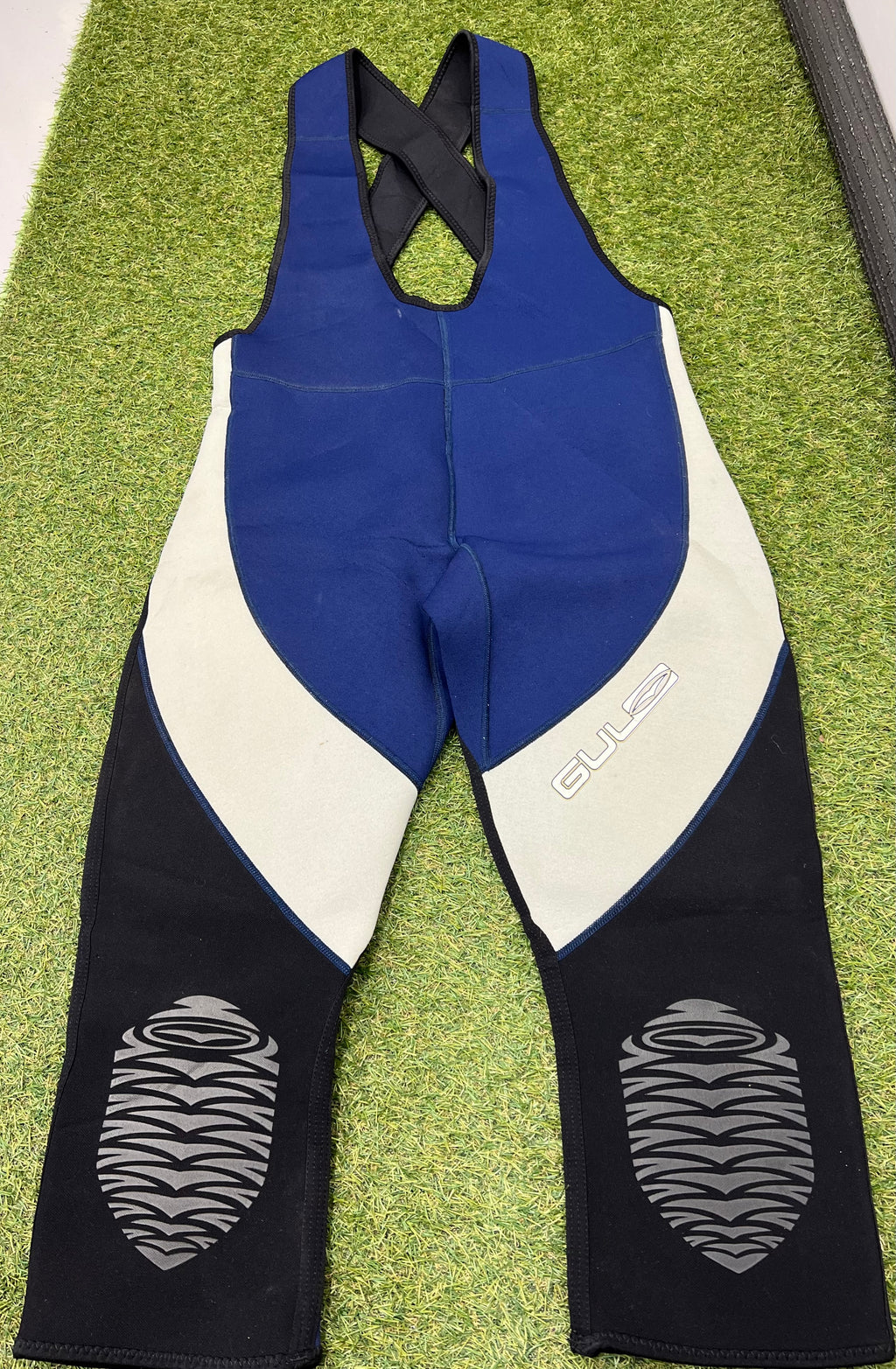 Front view of sleeveless blue, grey, and black neoprene wetsuit bib trousers with cross-back straps and padded knees, laid flat on grass