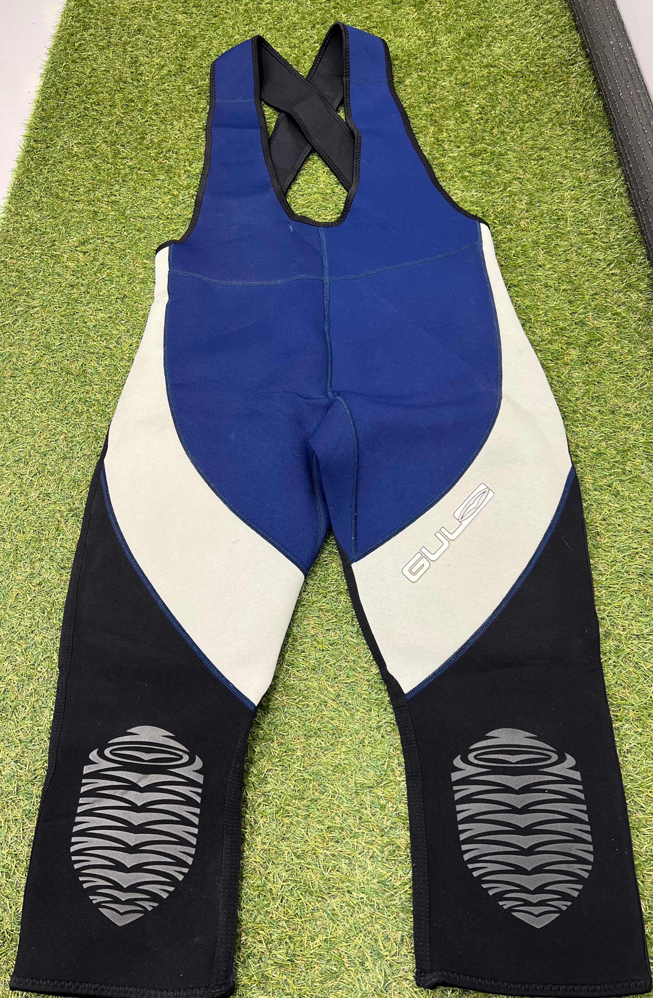 Front view of sleeveless blue, grey, and black neoprene wetsuit bib trousers with cross-back straps and padded knees, laid flat on grass