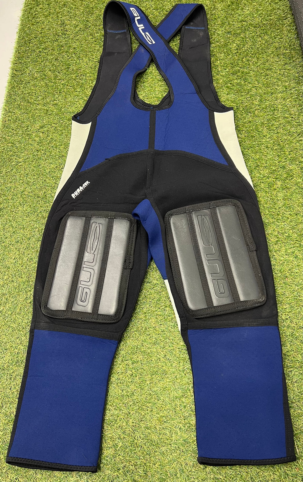 Back view of blue and black sleeveless wetsuit bib trousers showing thick padded knee protection panels with Gul branding, laid flat on grass