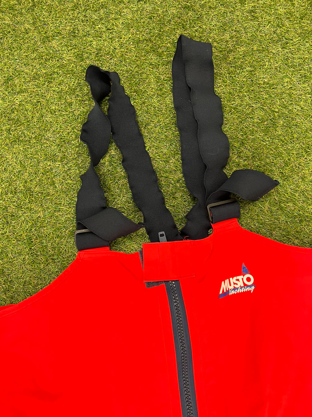 Top close-up showing black elastic shoulder braces and Musto logo above front zipper on red trousers.