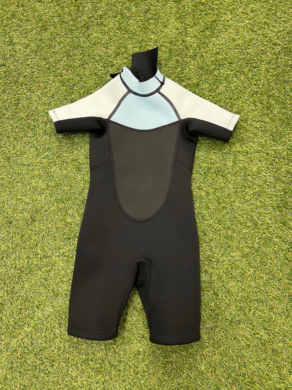 Front view of kids short sleeve wetsuit featuring black, light blue and white panels, laid flat on grass