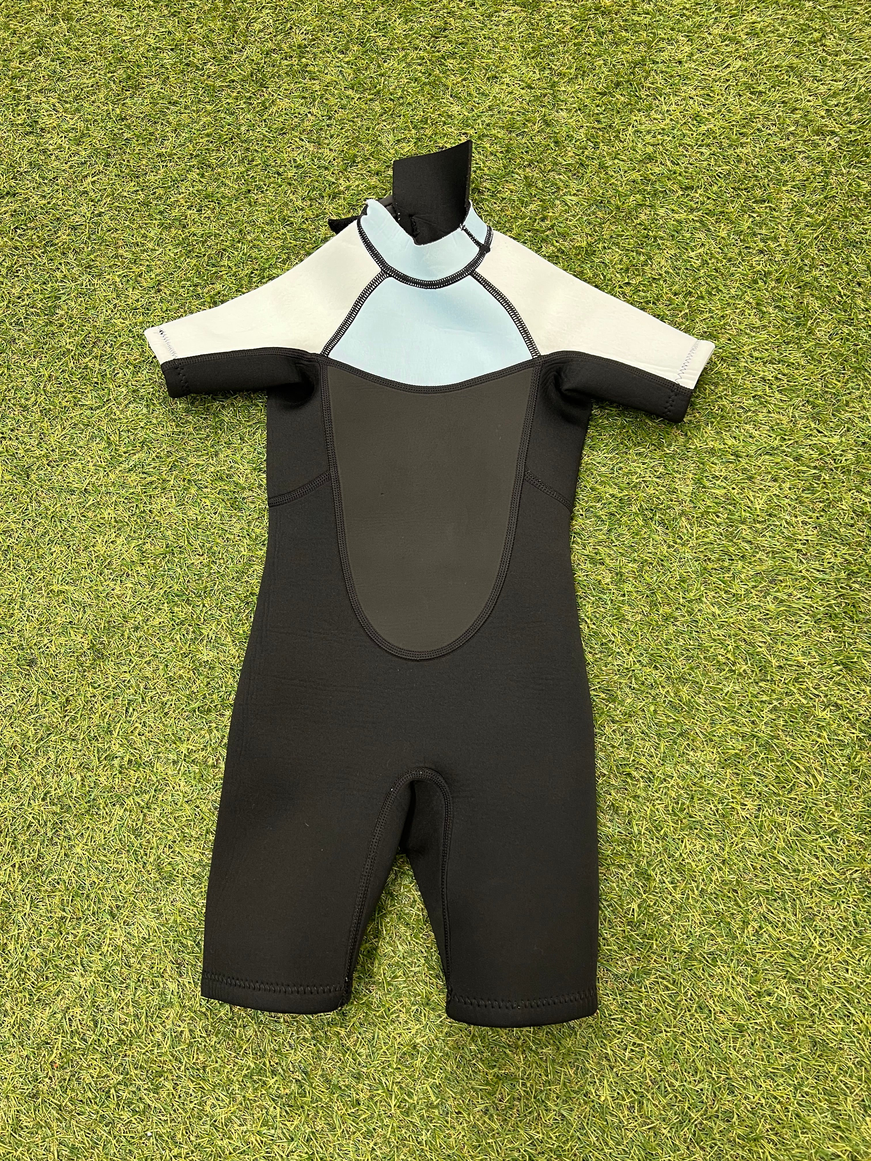 Front view of kids short sleeve wetsuit featuring black, light blue and white panels, laid flat on grass
