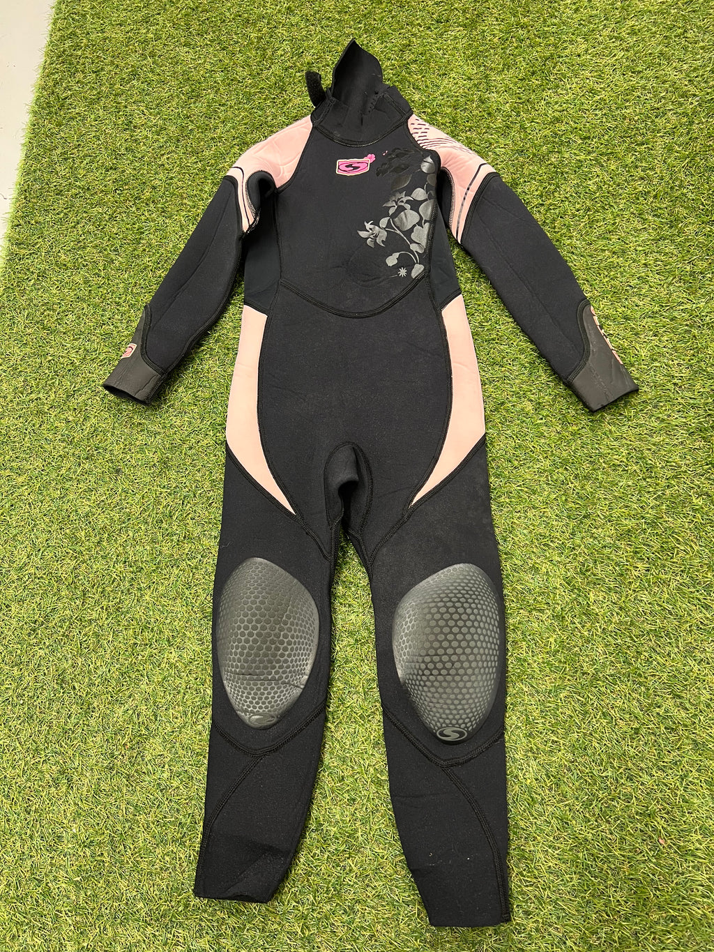 Front view of kids black and pink Sola wetsuit with floral detail and reinforced knee pads on green grass