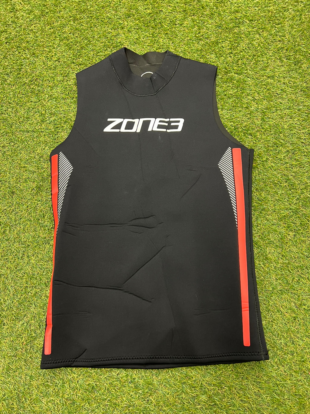 Front view of Zone3 black sleeveless wetsuit vest with red and white side detailing laid on grass