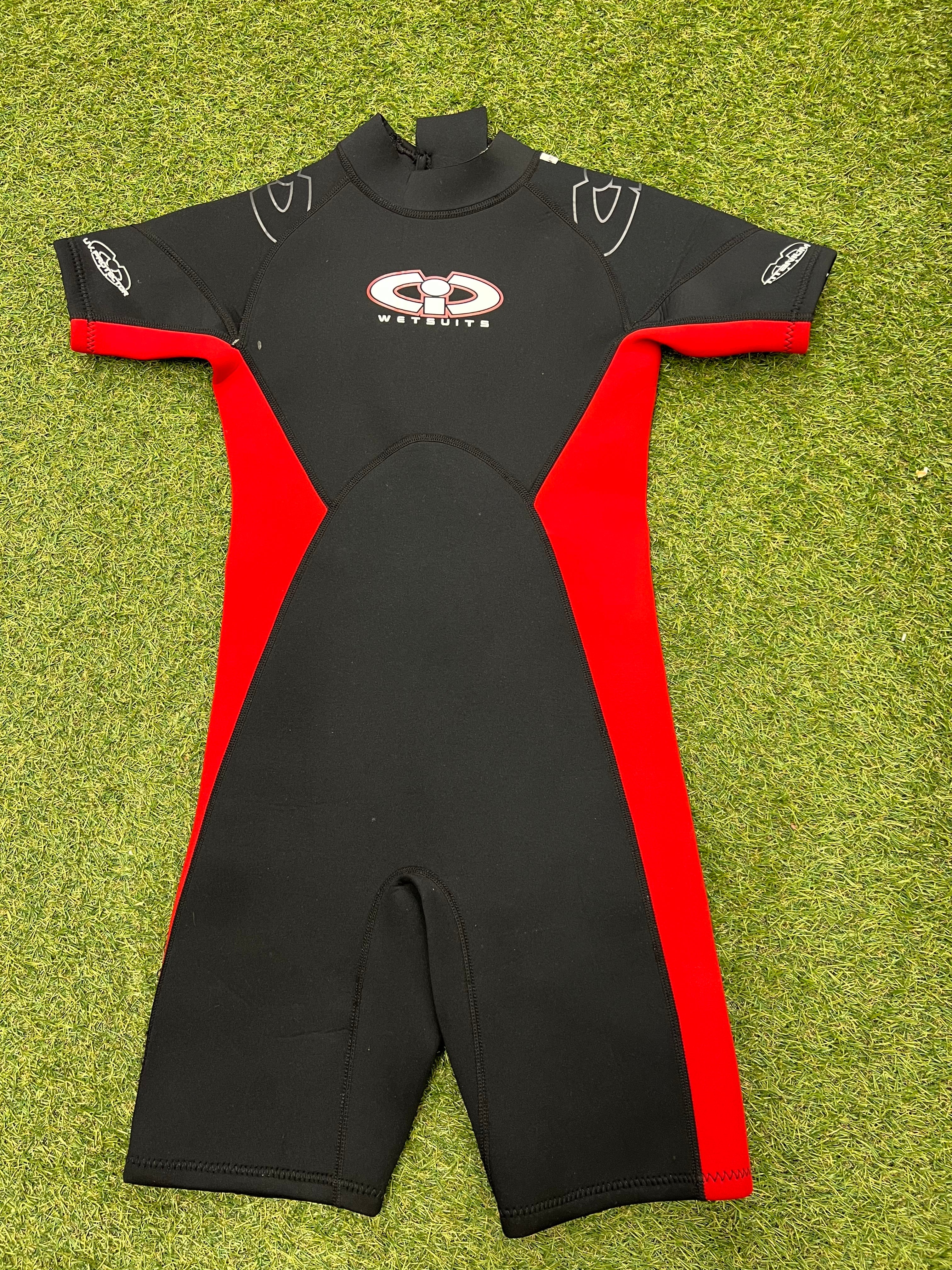 Front view of CIC kids shortie wetsuit in black with red side panels laid flat on grass