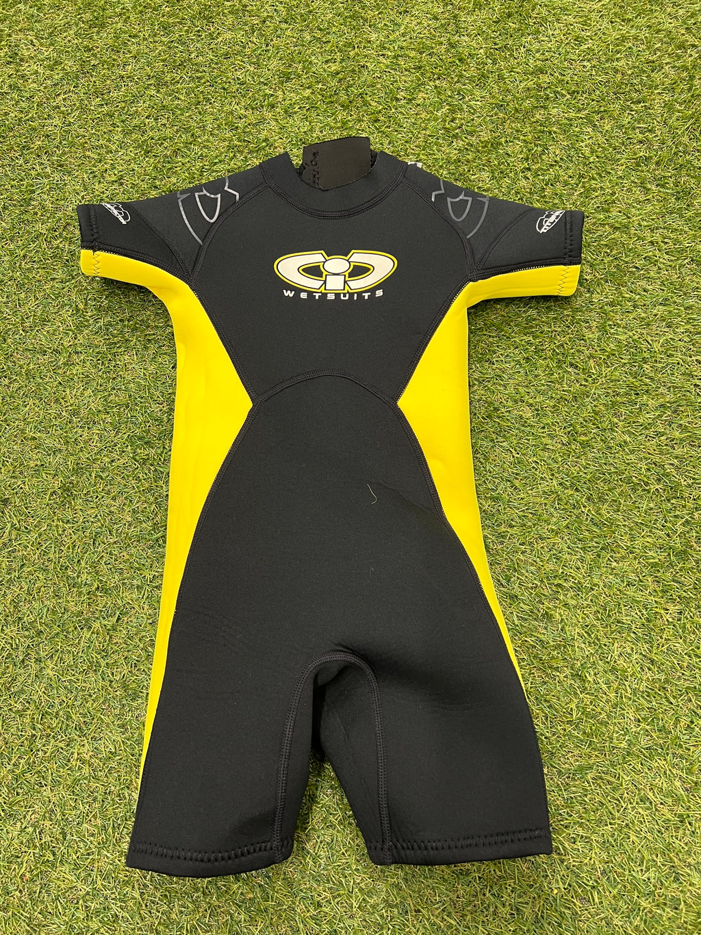 Front view of a black and yellow kids shortie wetsuit laid flat on grass, showing soft neoprene material and short sleeves