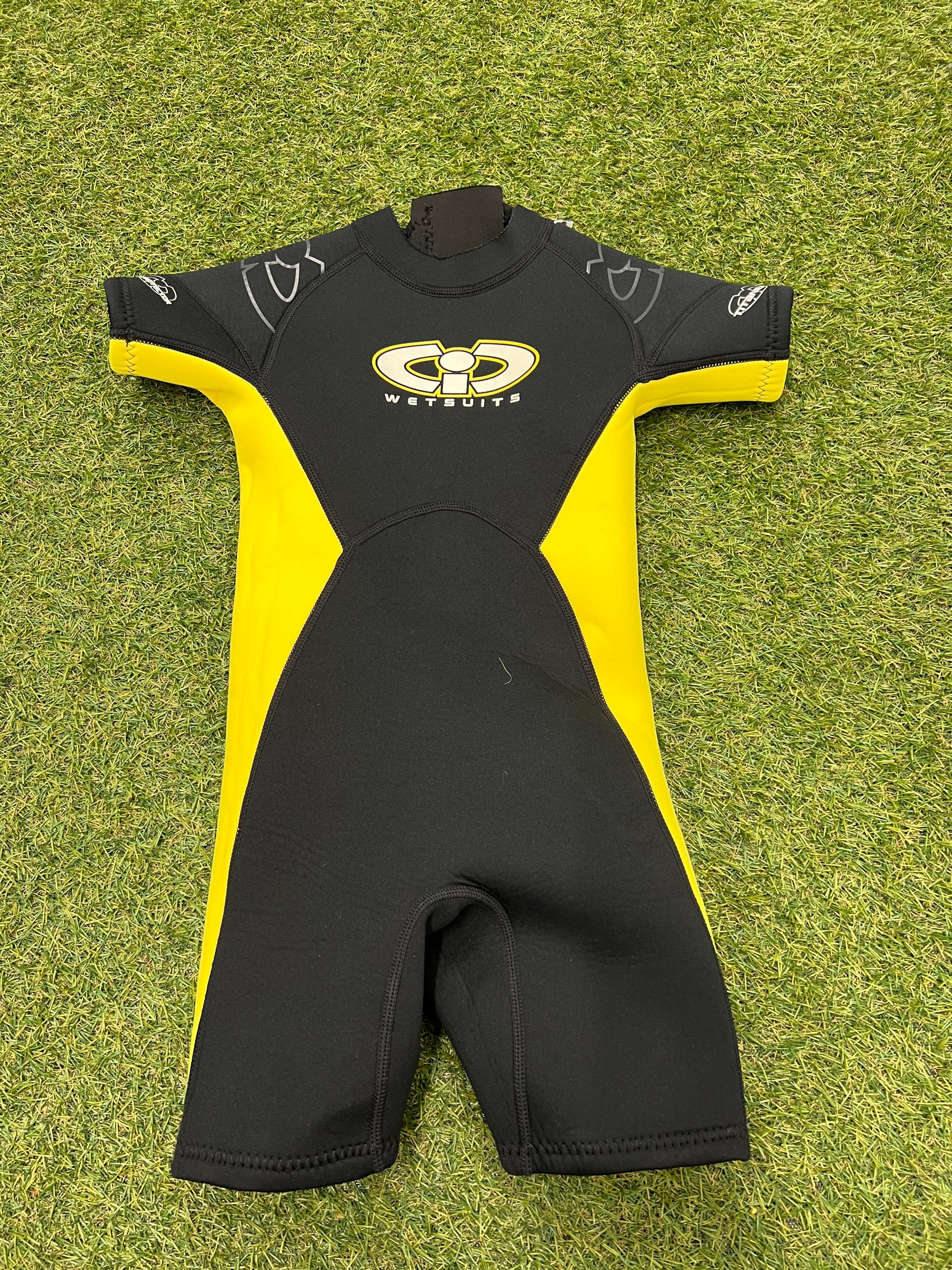 Front view of a black and yellow kids shortie wetsuit laid flat on grass, showing soft neoprene material and short sleeves