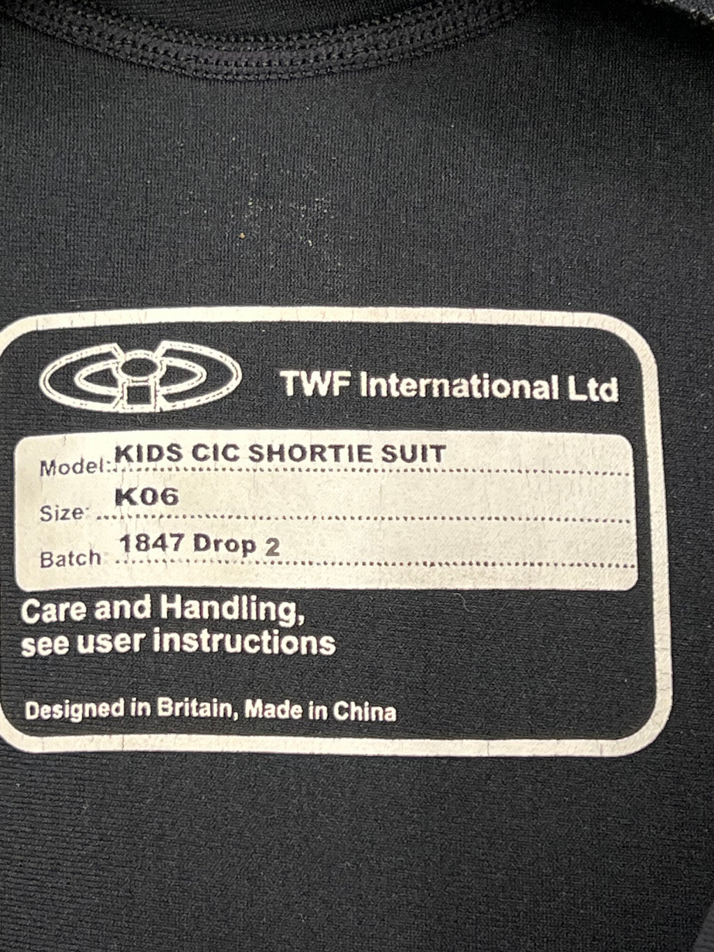 Close-up image of the inside label on a kids shortie wetsuit, showing brand TWF International Ltd, model Kids CIC Shortie Suit, size K06, designed in Britain, made in China