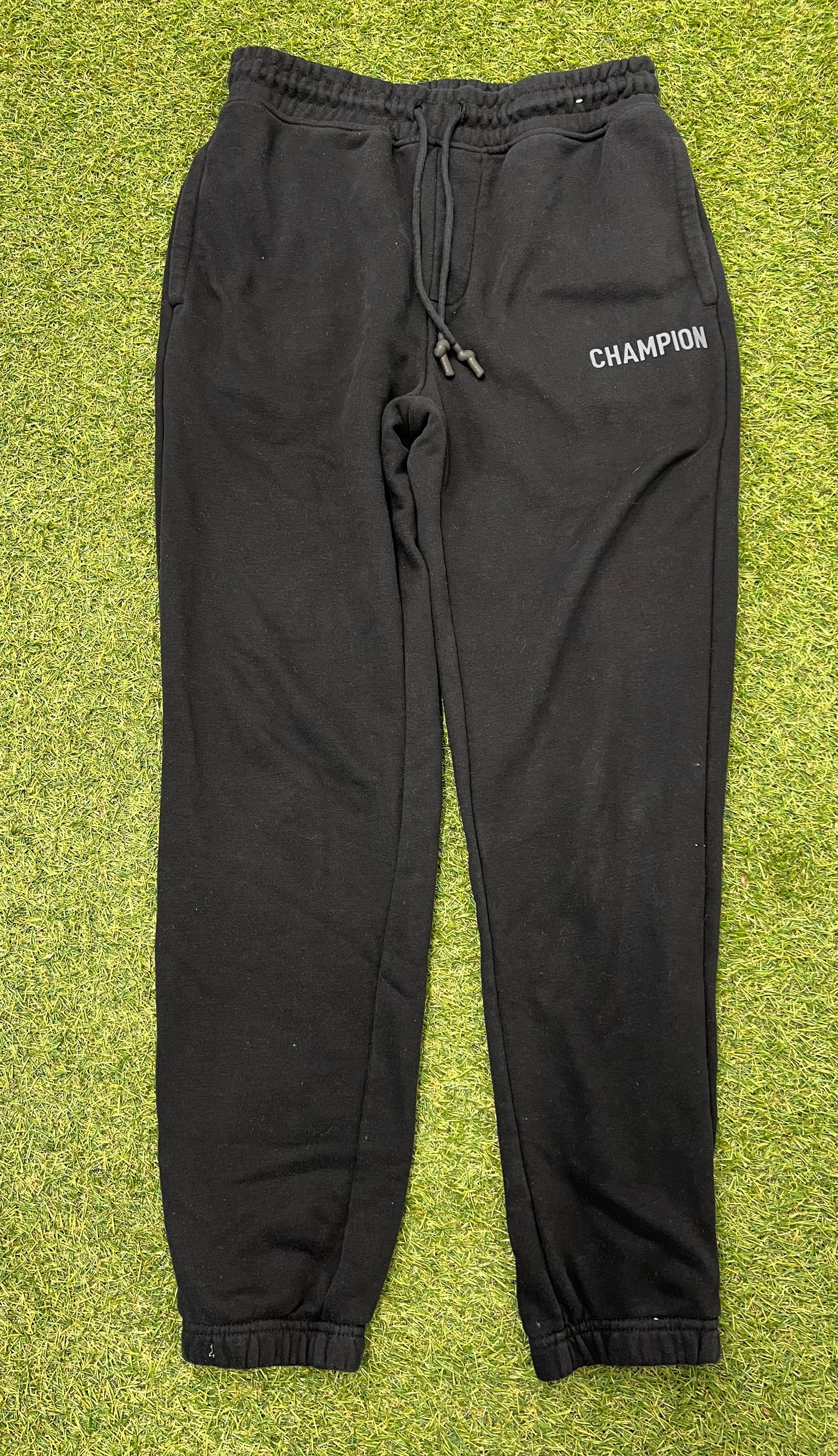 Front view of black Champion fleece tracksuit bottoms laid flat on grass, showing elastic waistband and drawstring
