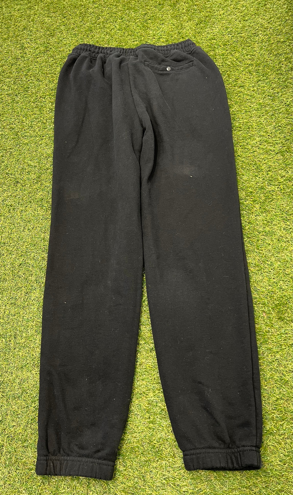 Back view of black Champion fleece tracksuit bottoms laid flat on grass, displaying rear pocket with button closure