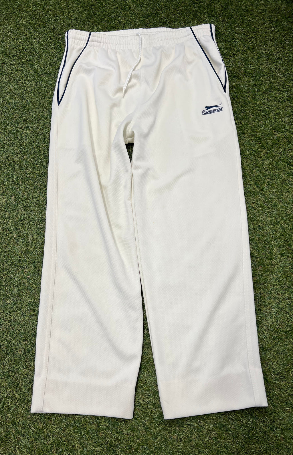 Front view of white youth Slazenger cricket trousers with navy piping on pockets and drawstring waist