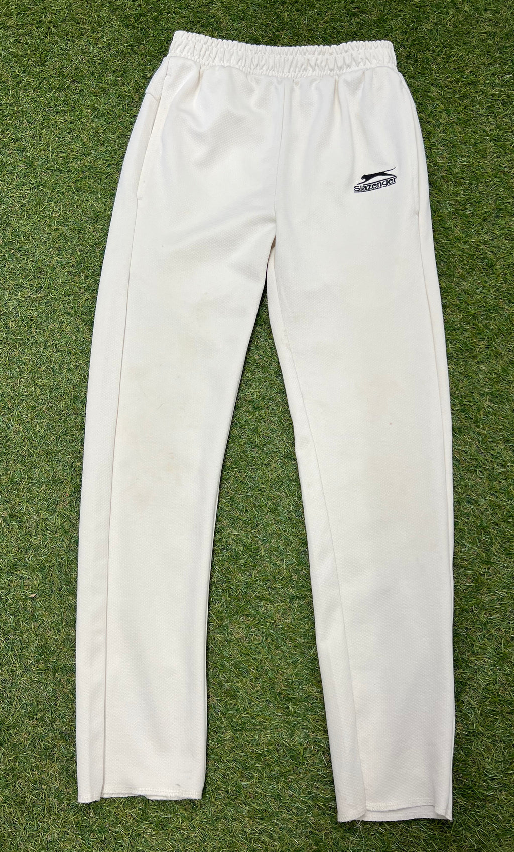 Front view of Slazenger kids white cricket trousers laid flat on grass