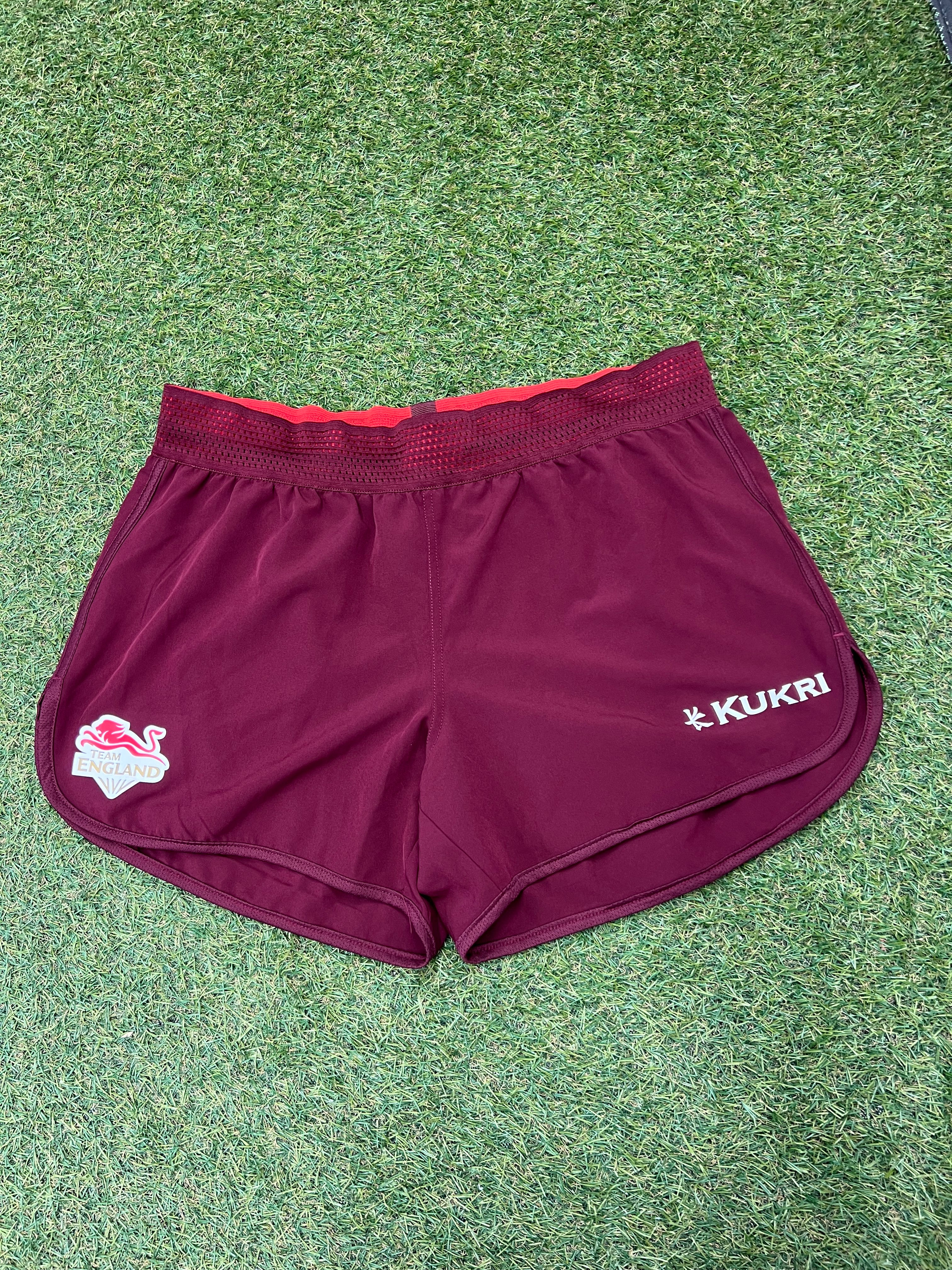 Front view of maroon Kukri Team England women's lightweight sports shorts with red mesh waistband on grass surface