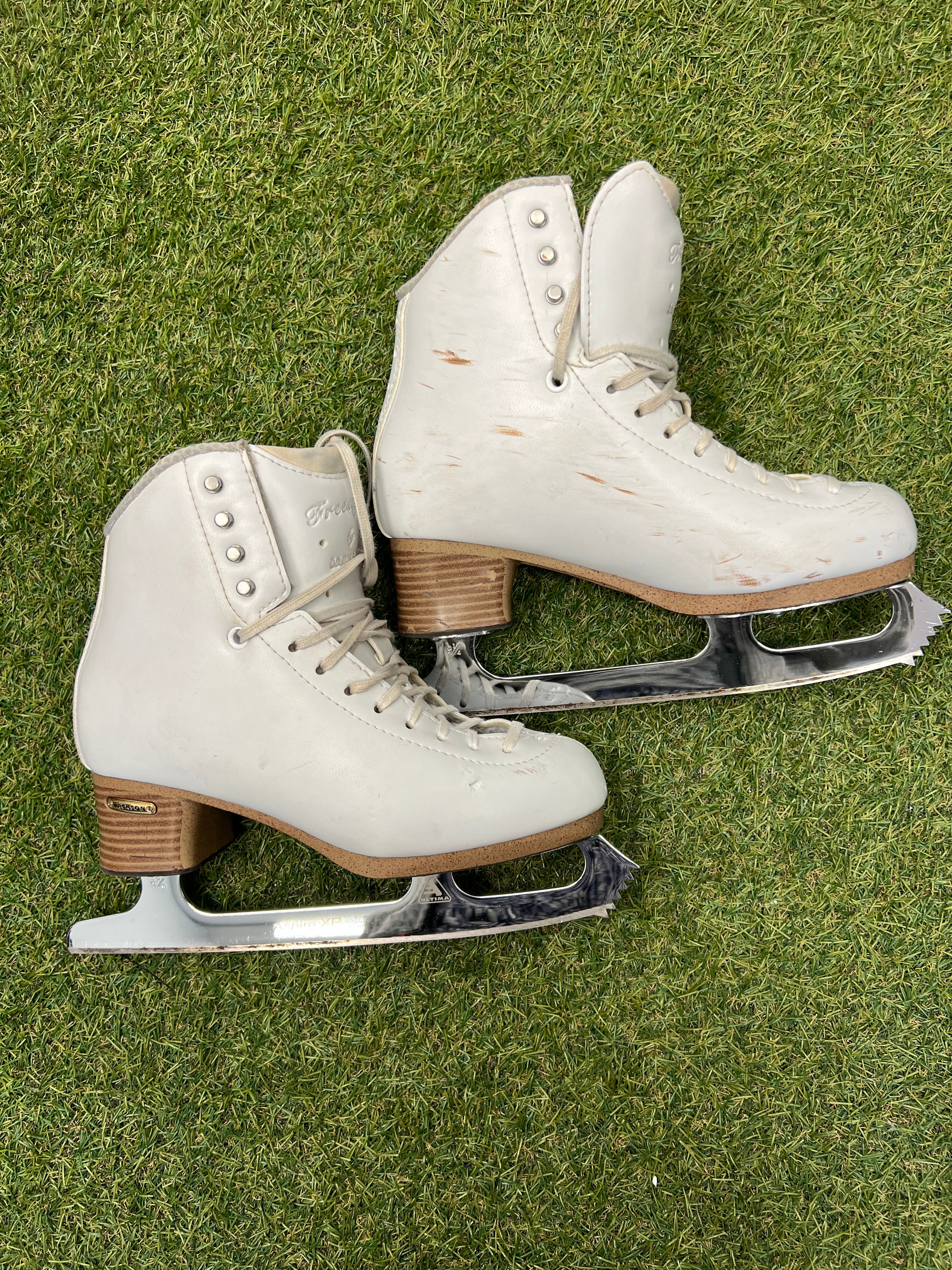 Pair of women’s white leather figure skates with stacked brown heels and chrome Aspire XP blades on grass