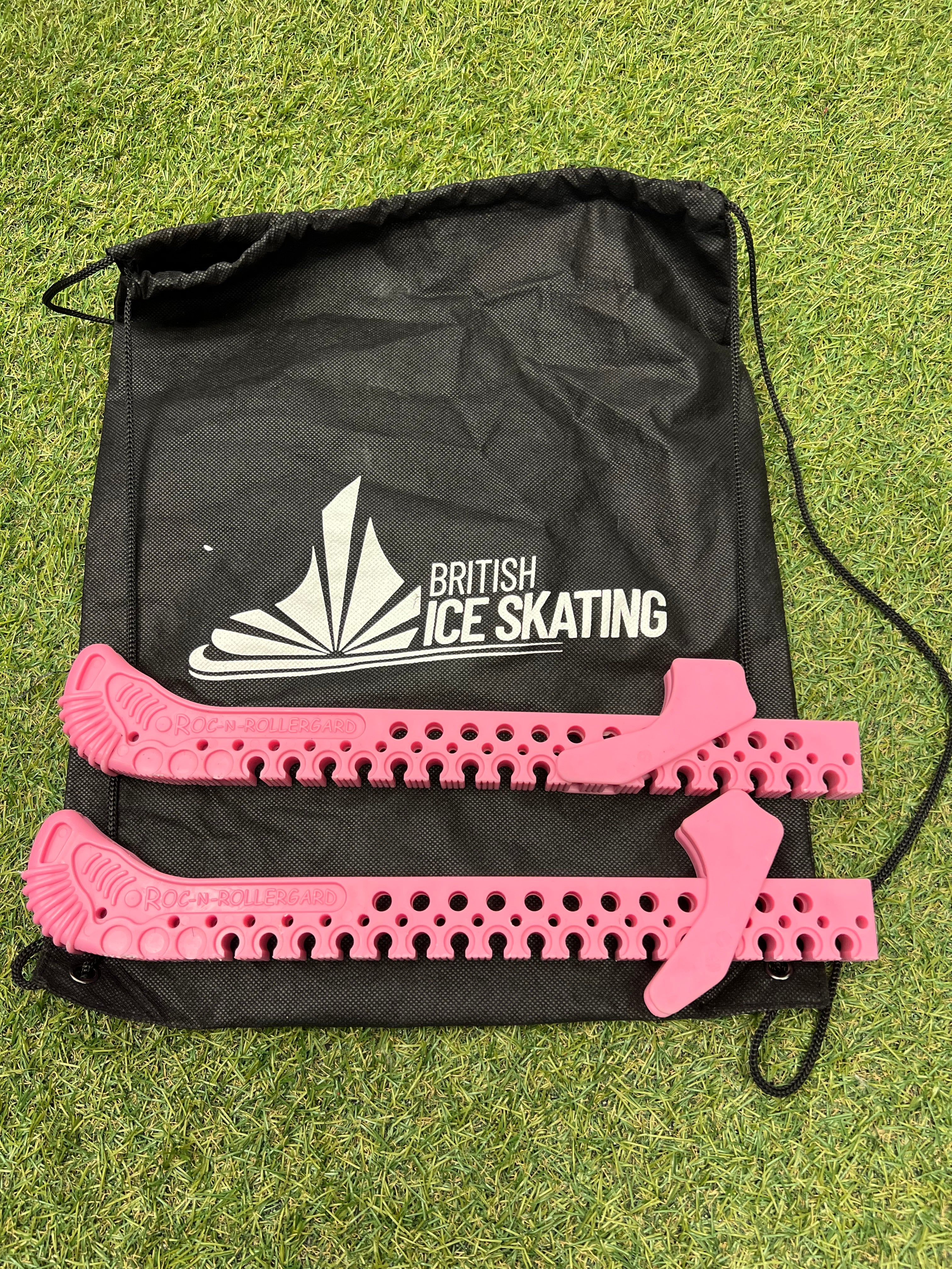 Pink blade guards and black British Ice Skating drawstring carry bag on grass surface