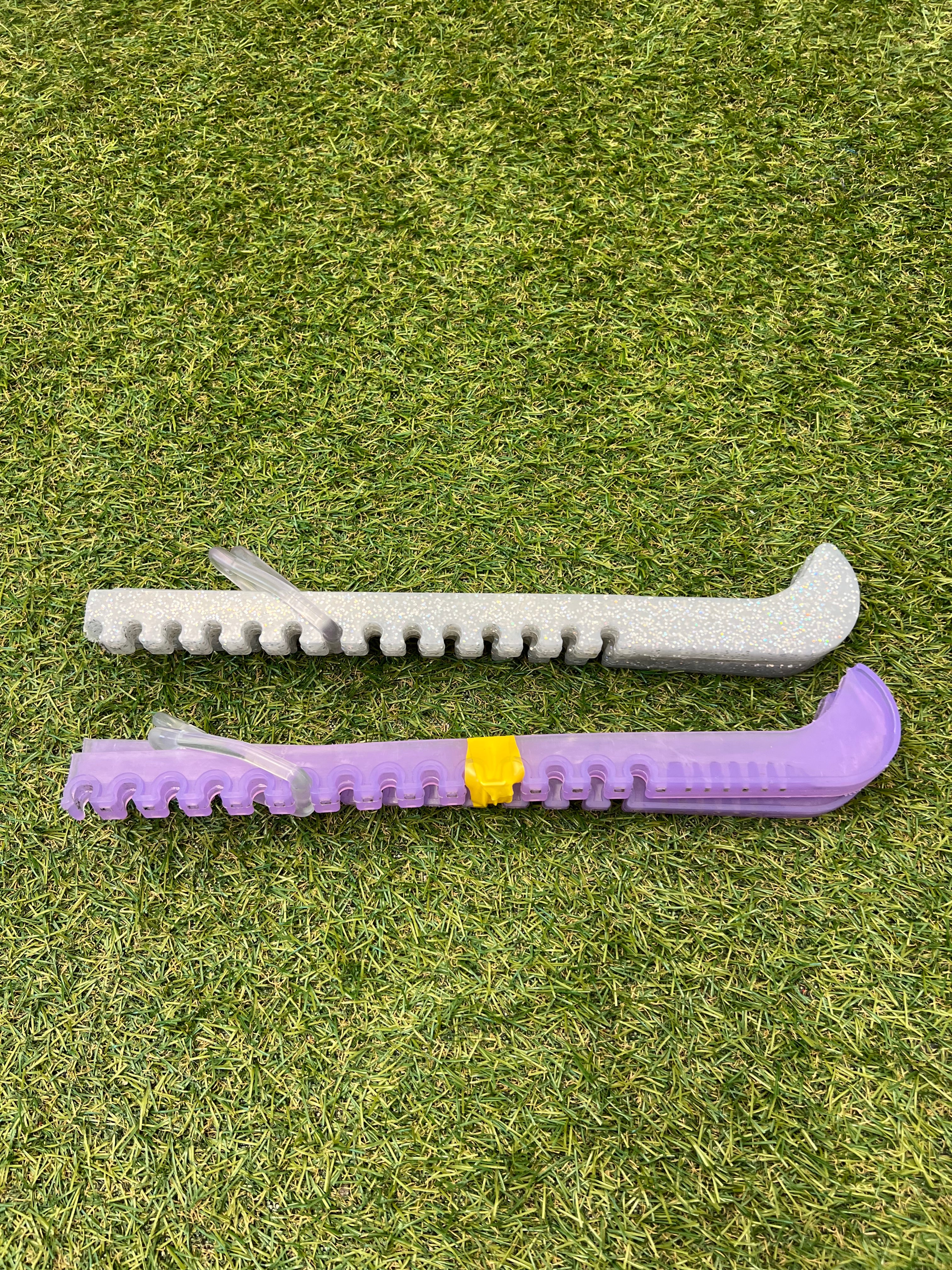 Silver and purple plastic ice skate guards with adjustable clips lying on green grass