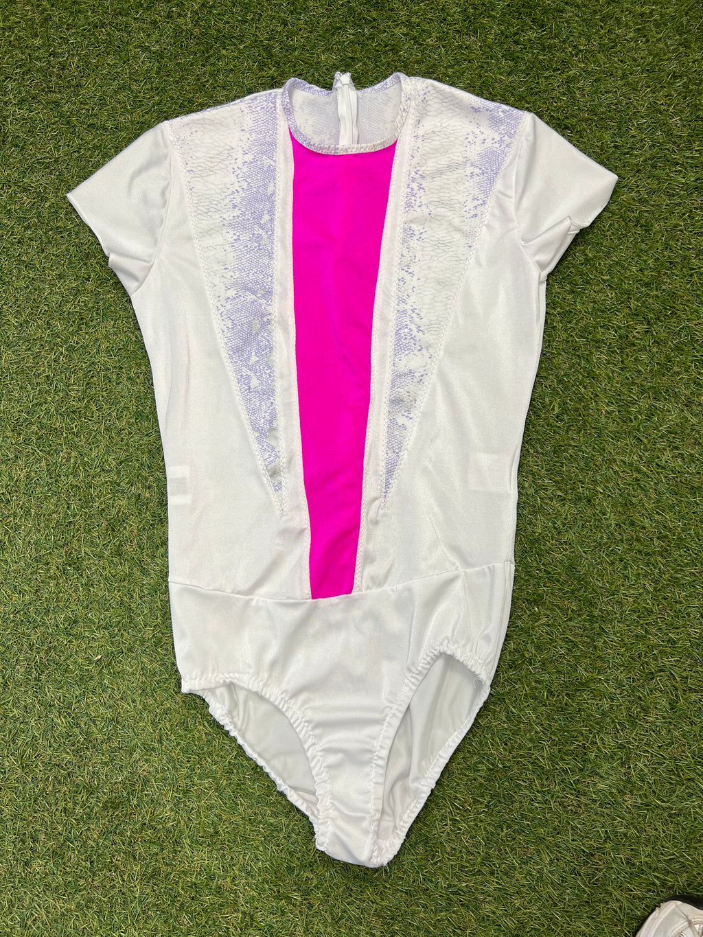 Front view of white short sleeve figure skating leotard with bright pink centre panel and lilac snake-print accents on grass background