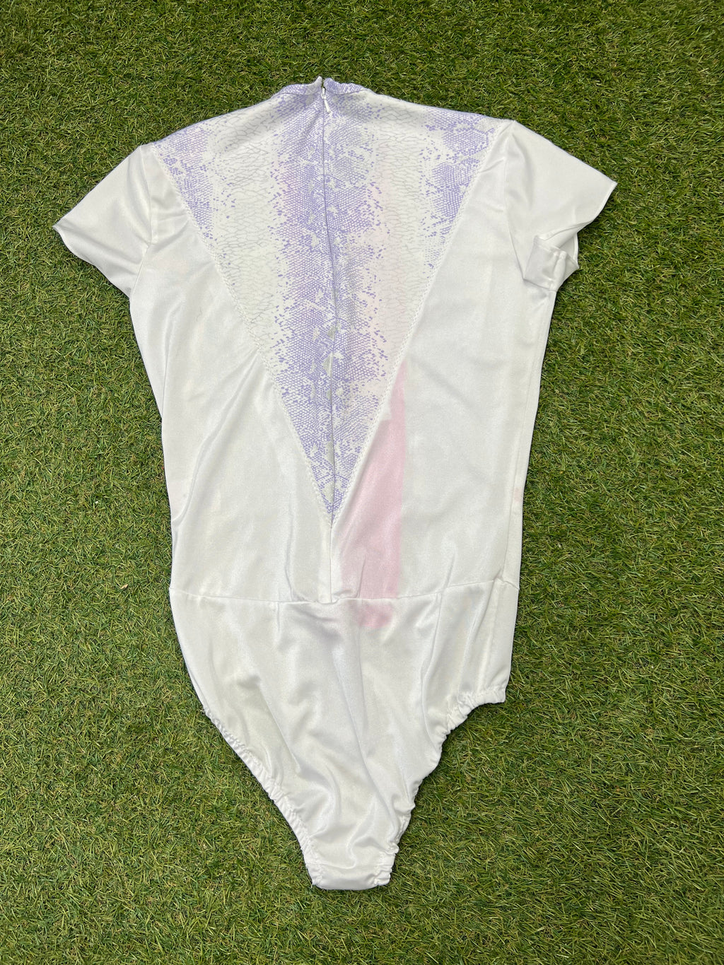 Back view of white short sleeve figure skating leotard with lilac snake-print detail and zip closure on grass background