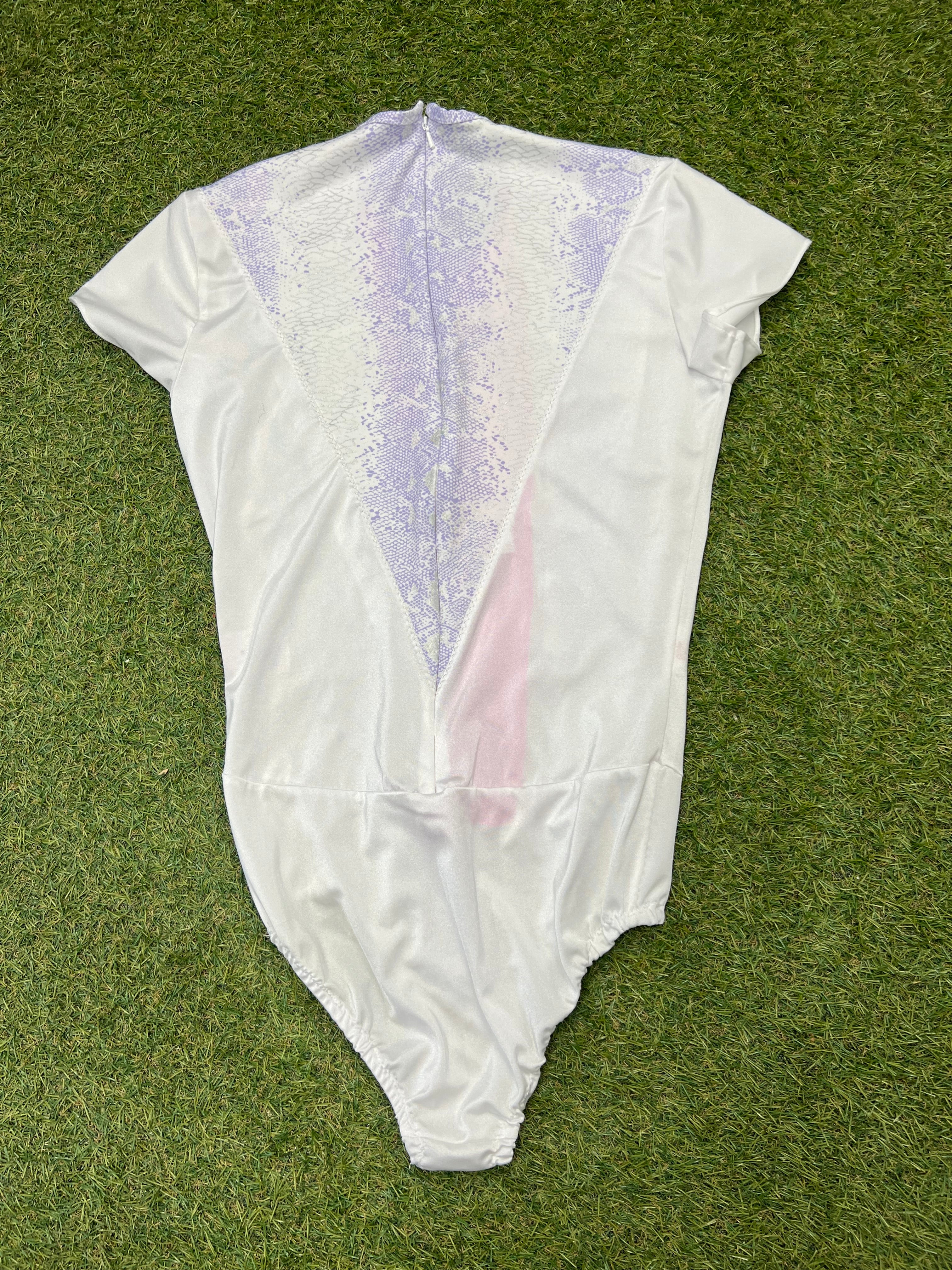Back view of white short sleeve figure skating leotard with lilac snake-print detail and zip closure on grass background