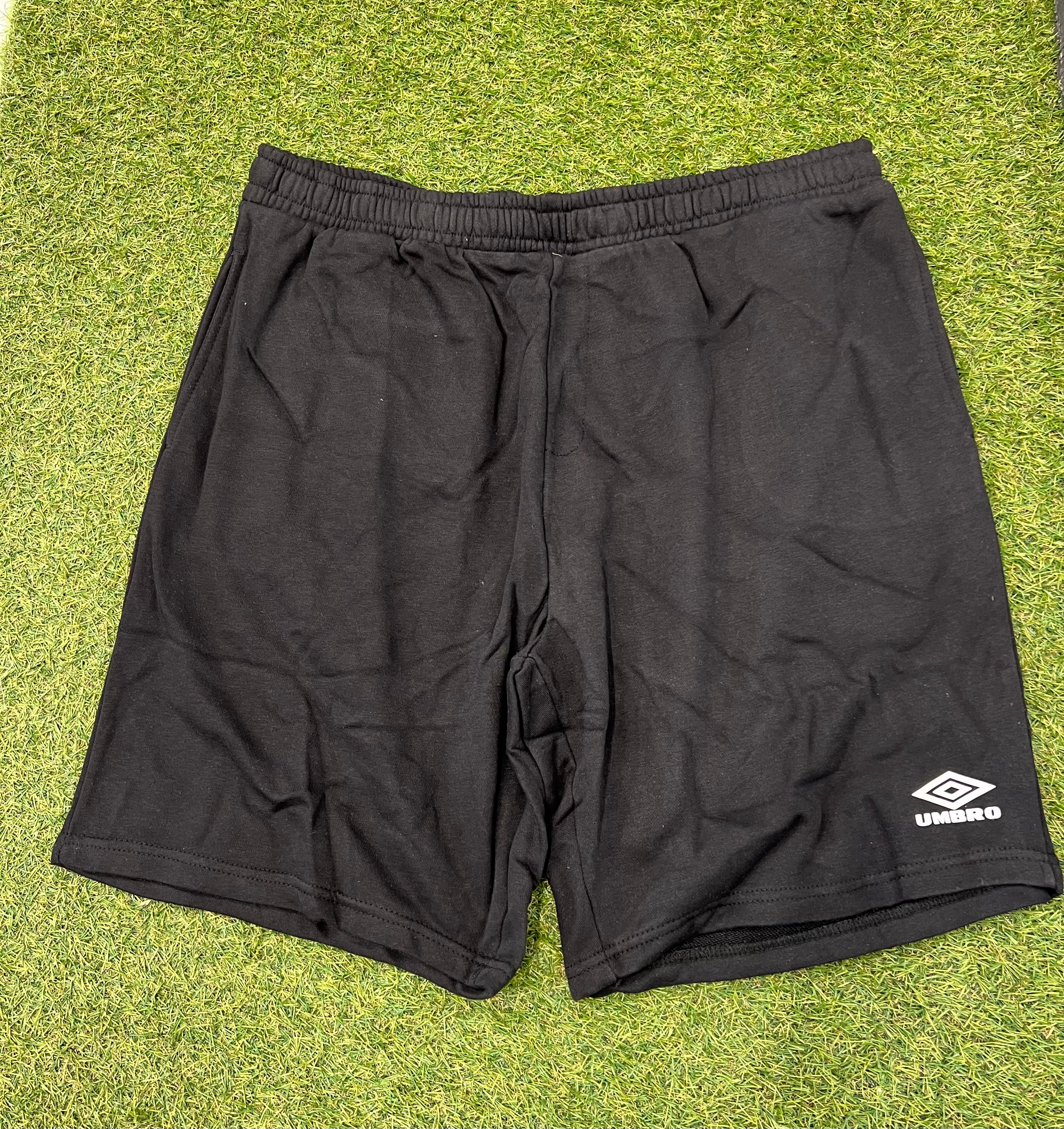 Front view of black Umbro Diamond Jog Shorts laid on grass showing elastic waistband and logo