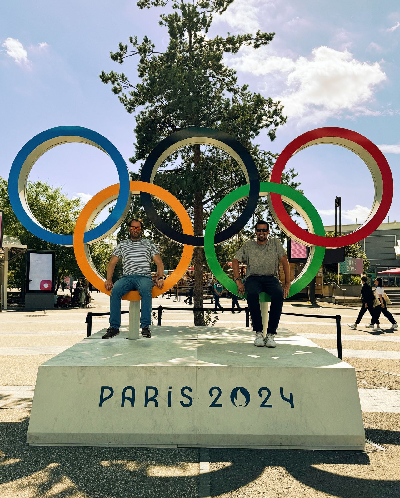 Two people sitting on Olympic rings with 'Paris 2024' below, against a blue sky.