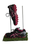 Pair of Asics black and pink hockey trainers displayed on artificial grass with logo backdrop