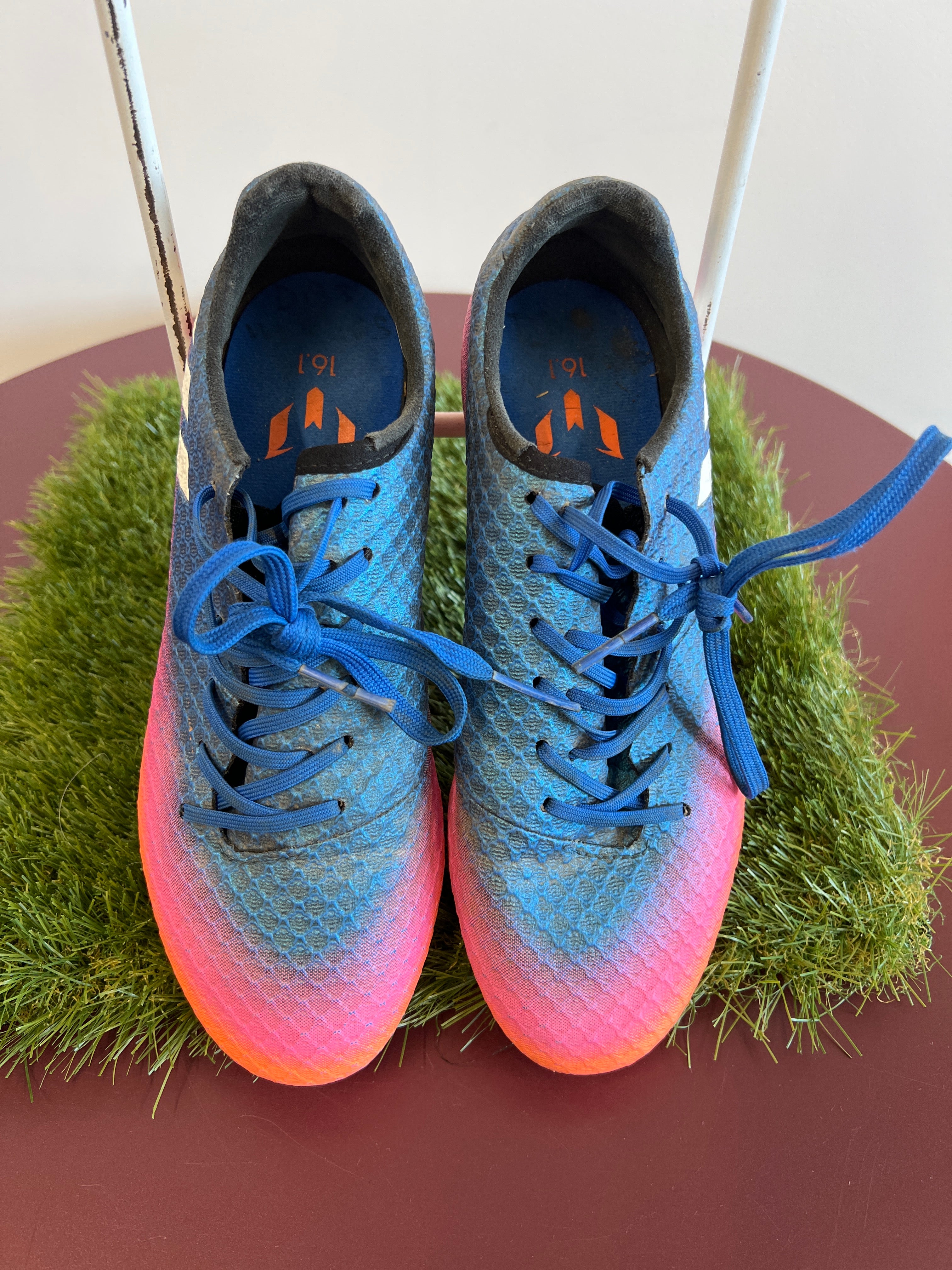 Top view of kids adidas football boots in blue, pink, and orange gradient with blue laces on artificial grass