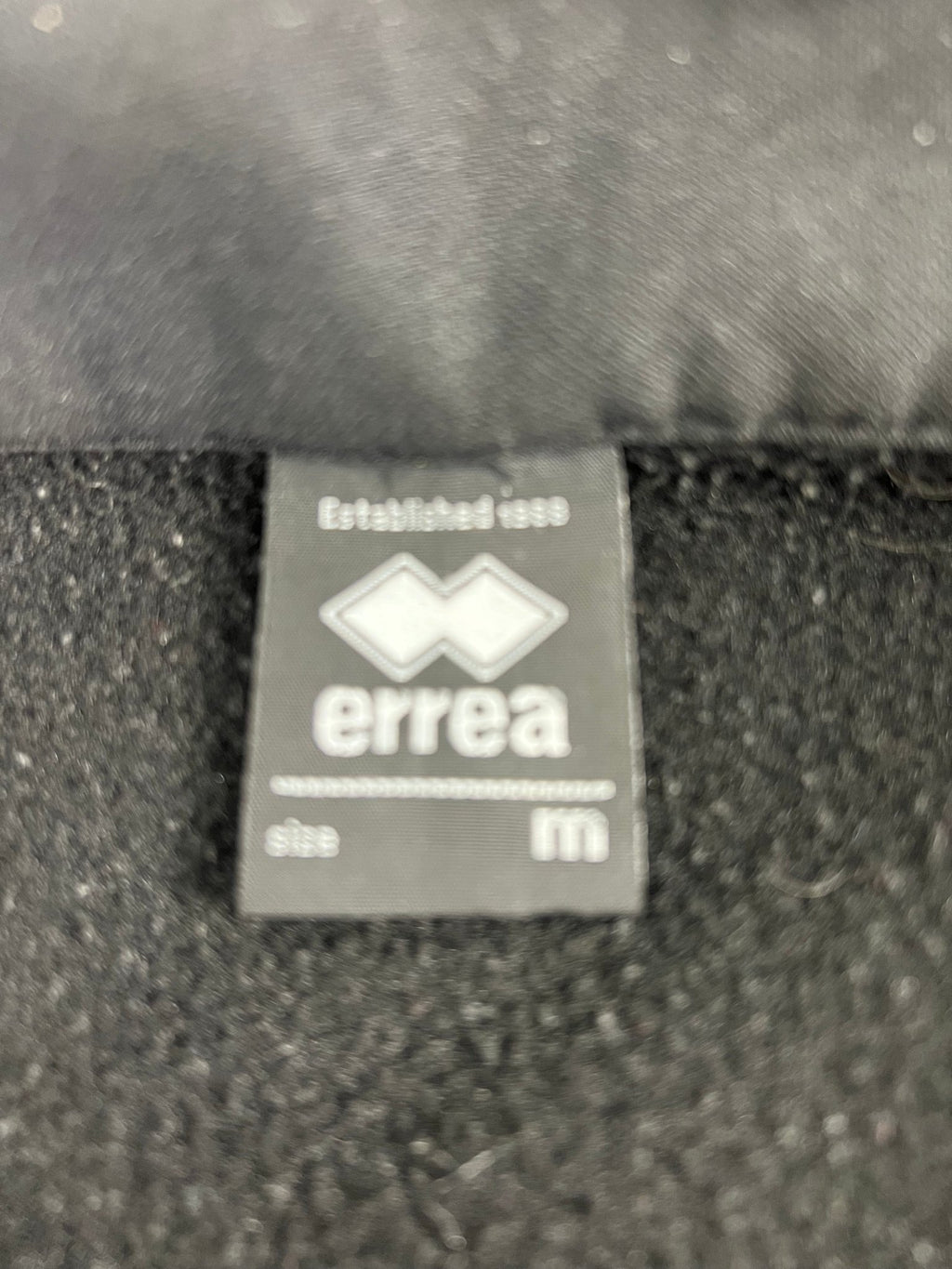 Close-up of errea brand label inside men’s black training top indicating size medium