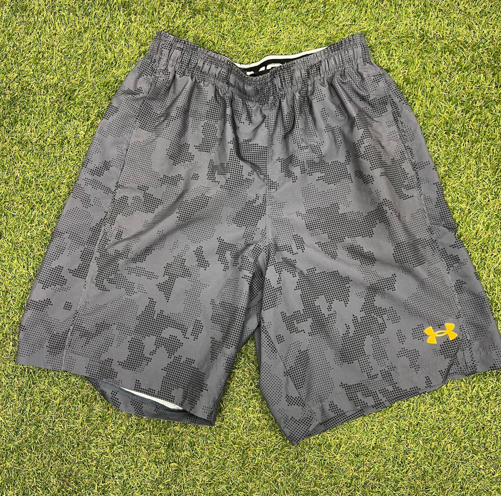 Front view of men's Under Armour grey camo loose fit shorts with yellow logo on grass background