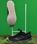 A pair of black Mizuno TC-01 trainers, one showing the sole and one in profile on artificial grass with a green background.