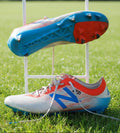 Side and sole view of New Balance Revlite football boots in white, blue, and red on grass display