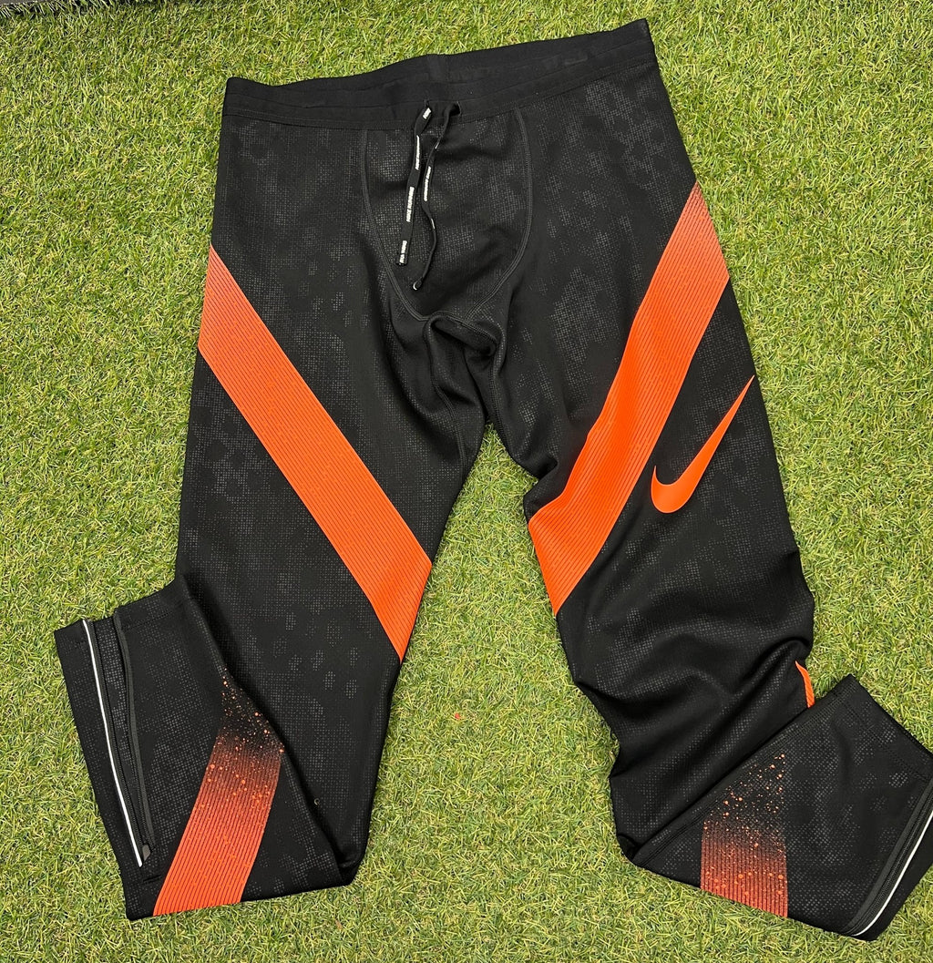 Front view of black and orange Nike Dri-FIT running tights with ankle zips displayed on grass