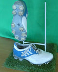 Pair of adidas white and blue golf shoes with spiked soles displayed on a grass mat