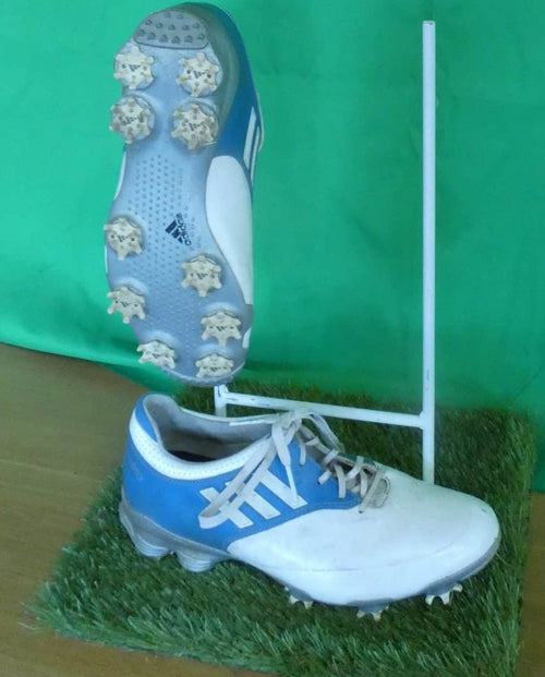 Pair of adidas white and blue golf shoes with spiked soles displayed on a grass mat